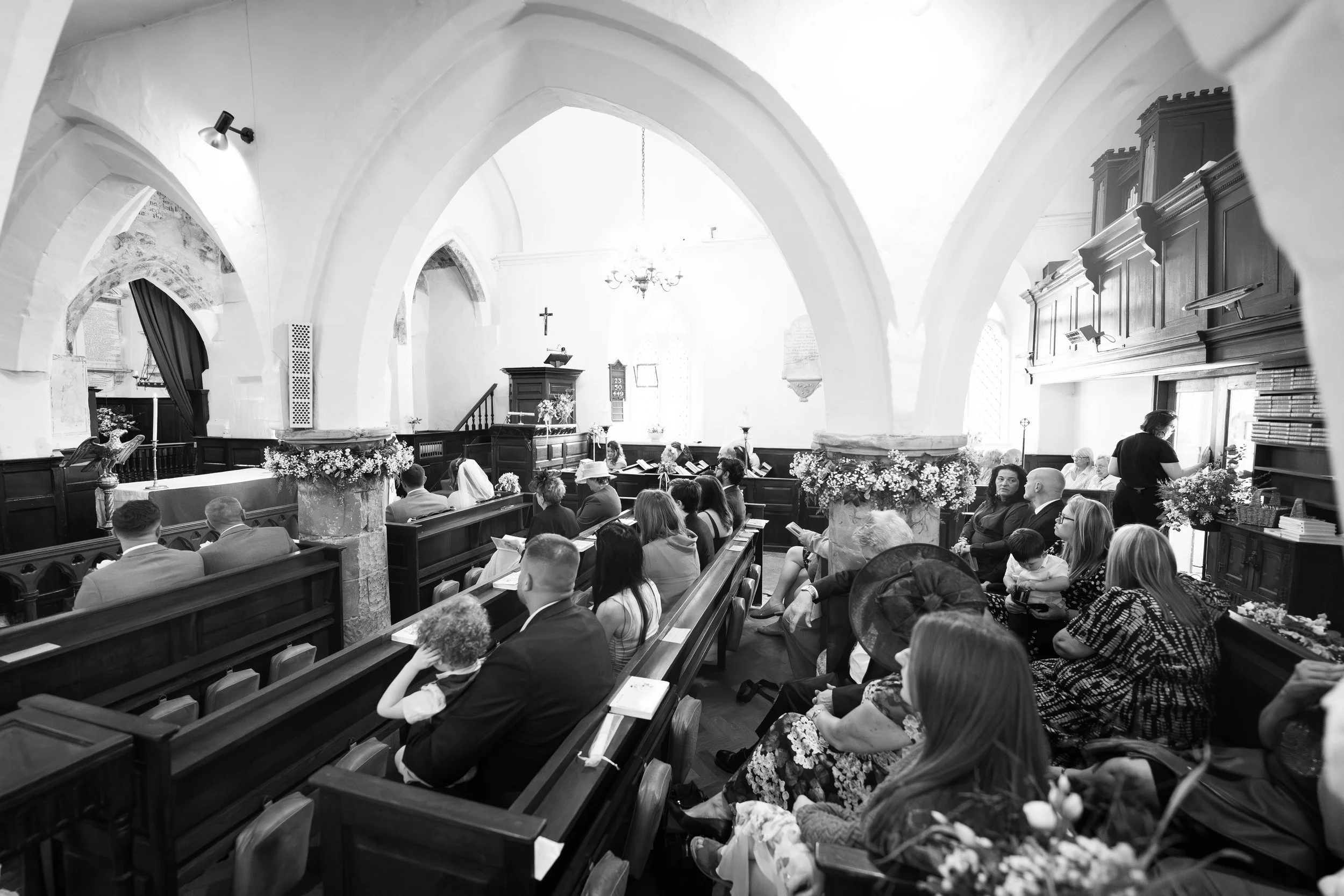 People seated in pews inside a church with arched ceilings, floral decorations on columns, and an altar at the front.