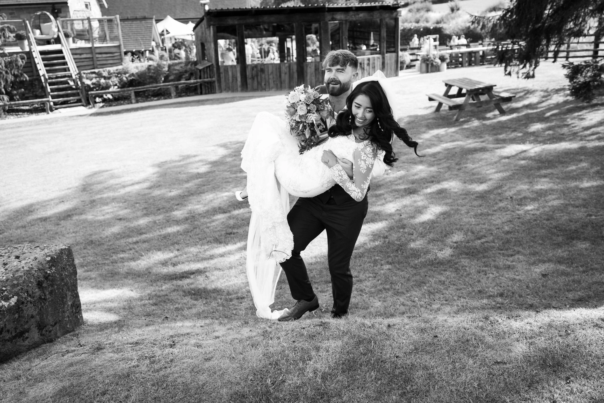 A groom carrying a bride in a white lace wedding dress and veil on a grassy outdoor area with picnic tables and wooden structures in the background, smiling and enjoying the moment.