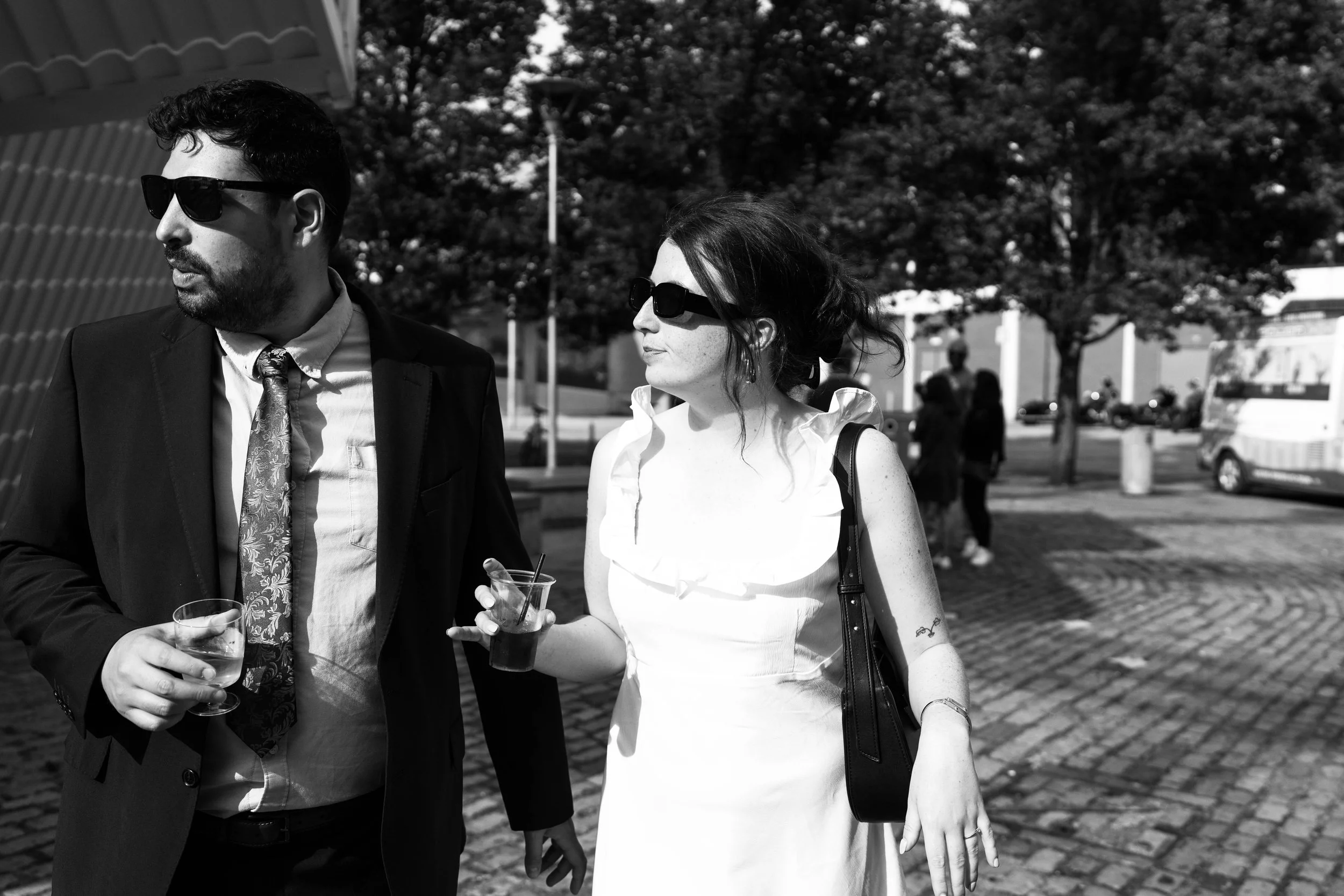 Two people, a man and a woman, walking outdoors in daytime, both wearing sunglasses and carrying drinks, with trees and parked vehicles in the background.