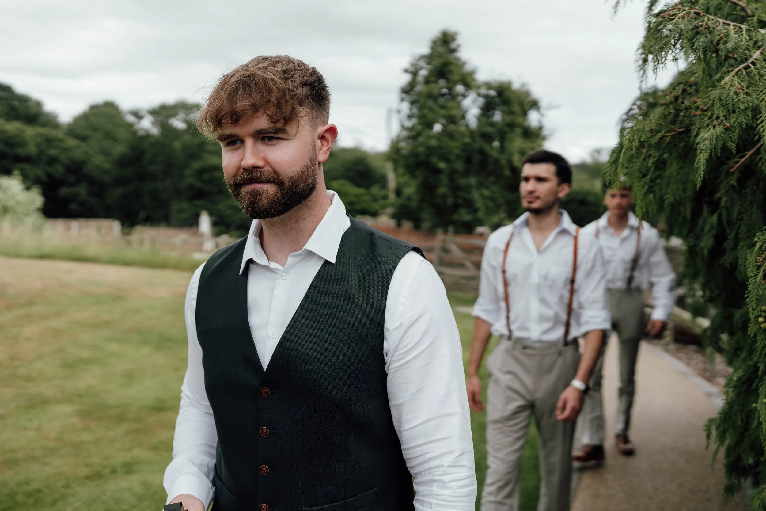Three men outdoors on a cloudy day, dressed in vintage-style clothing, with one in the foreground wearing a vest and white shirt, and two others in the background walking along a dirt path, surrounded by greenery.