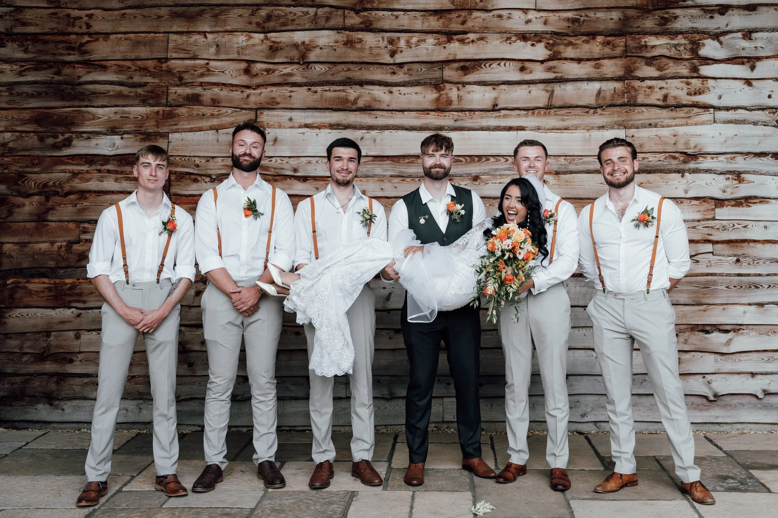 A bride in a white wedding dress being held by a groom in a black vest and pants, surrounded by six groomsmen in white shirts, beige trousers, and orange suspenders, against a wooden plank wall.