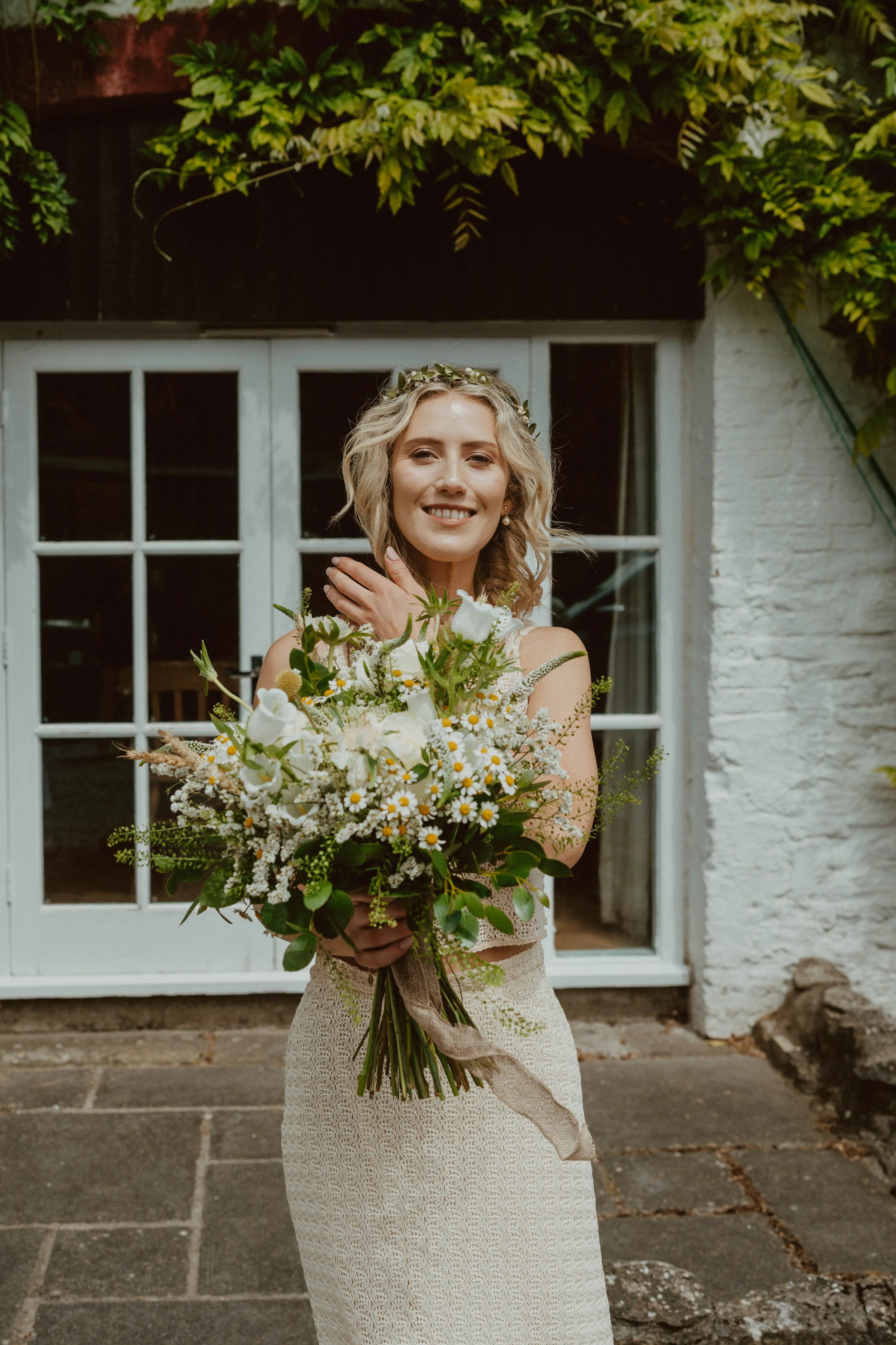 A smiling woman in a lace dress, holding a big bouquet of white and green flowers. She is wearing a flower crown and standing outside in front of a white door with glass panels.