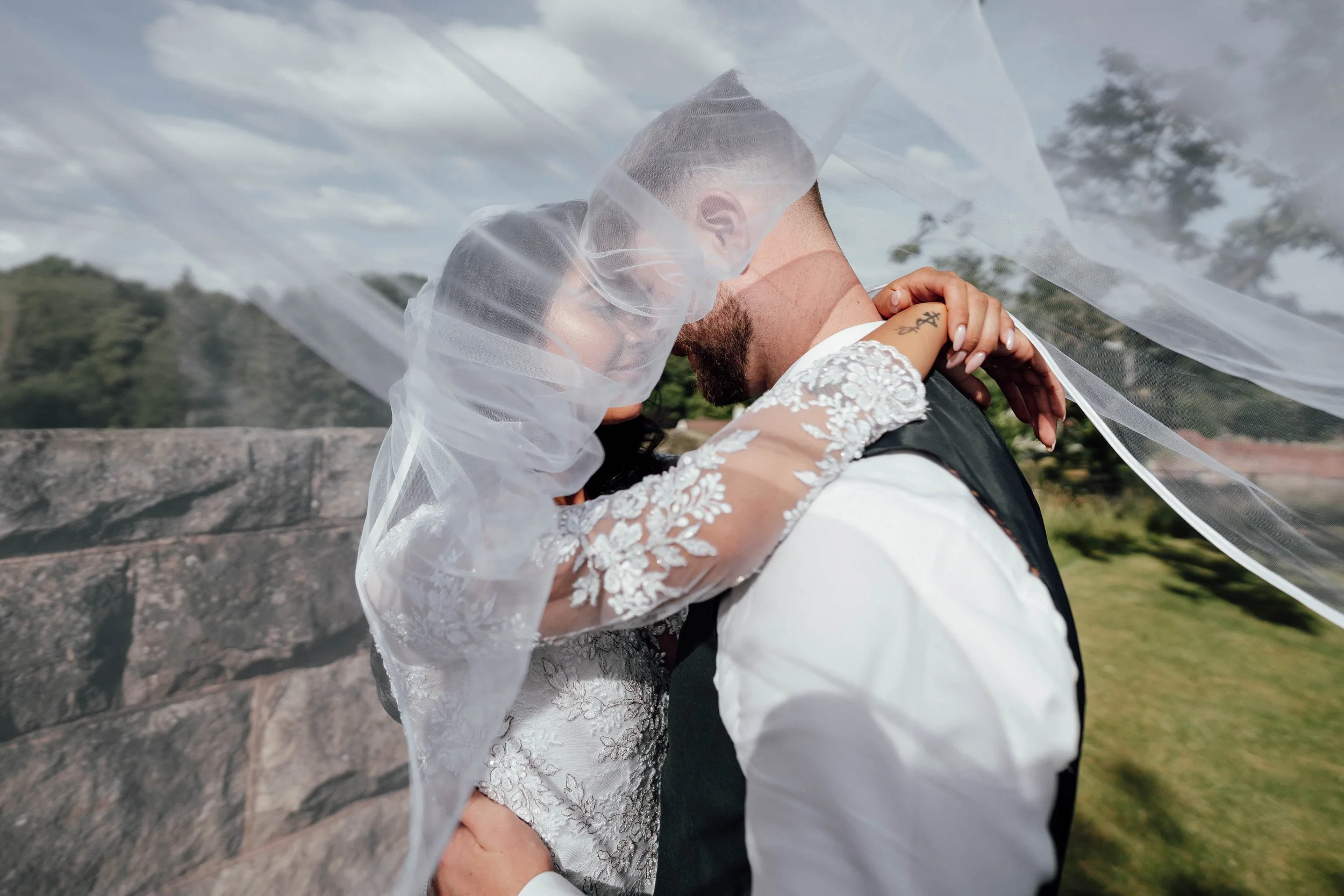 A newlywed couple sharing an intimate moment outdoors, with the bride wearing a lace wedding dress and veil, and the groom in a white shirt and vest, embracing closely beside a stone wall with greenery and trees in the background.