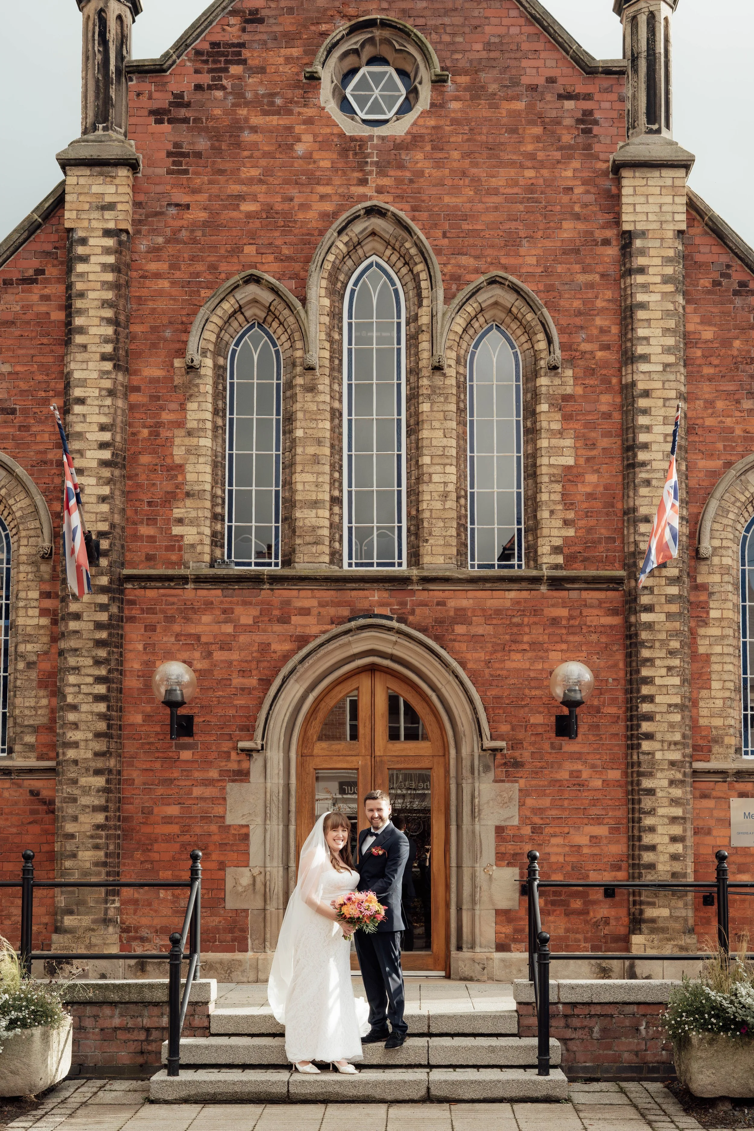 Bride and groom standing in front of a brick church, smiling and holding a bouquet, celebrating their wedding.