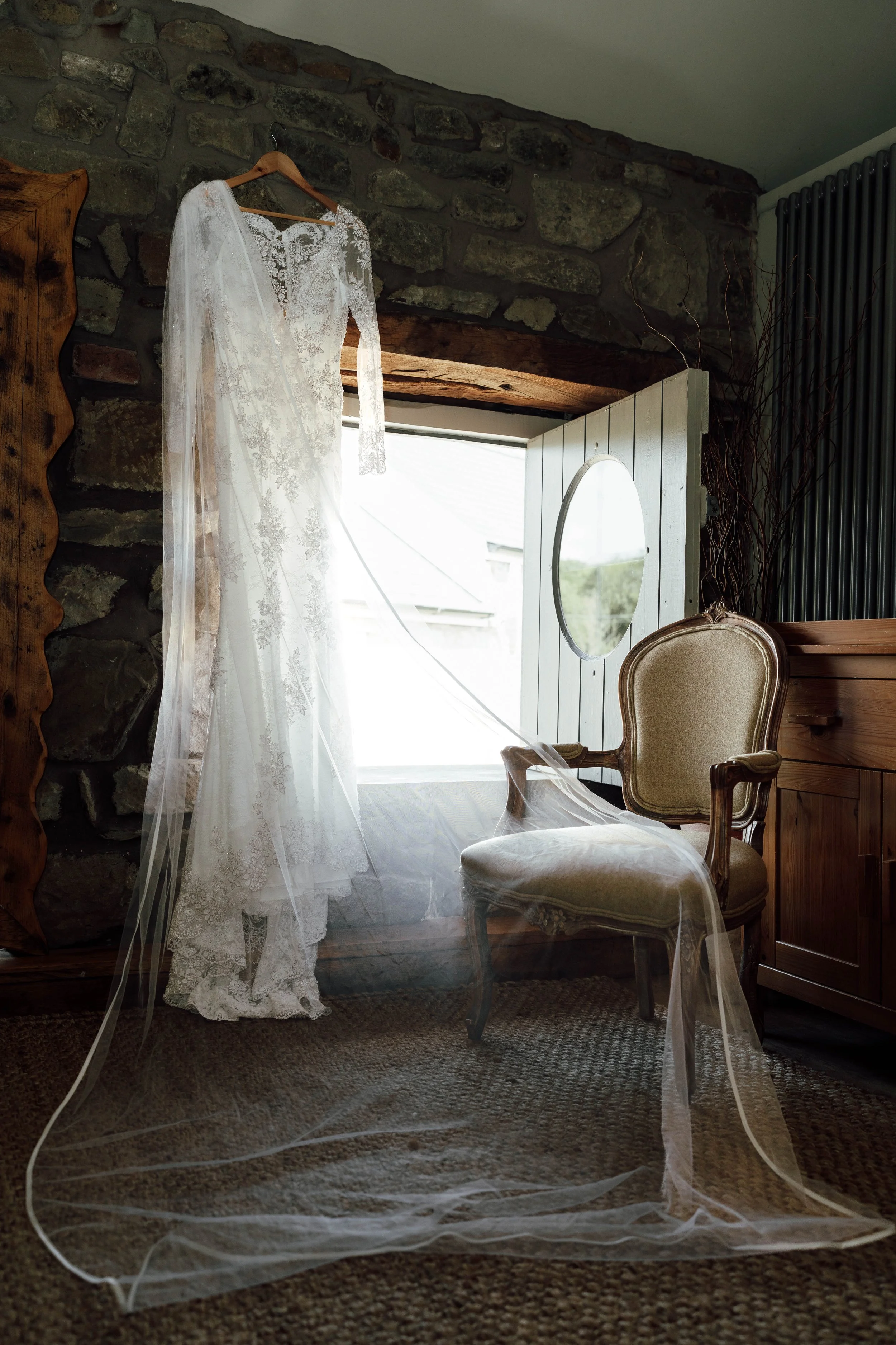 A white lace wedding dress hanging on a hanger in a rustic room with a stone wall, an antique chair, and a mirror