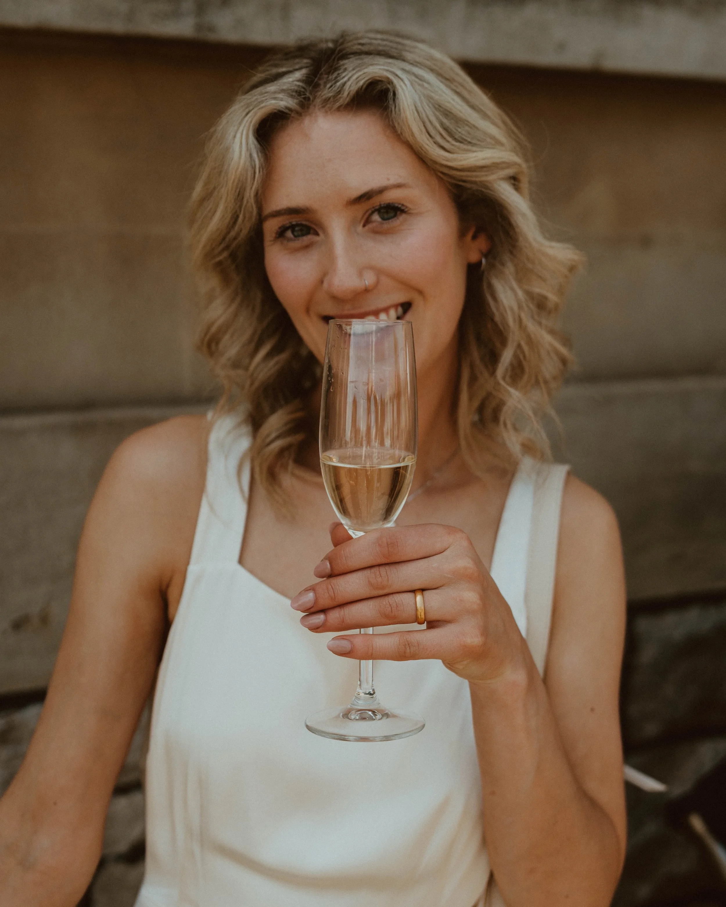 A woman with blonde, wavy hair and a nose piercing is holding a glass of champagne, smiling, and wearing a white sleeveless top in an indoor setting.