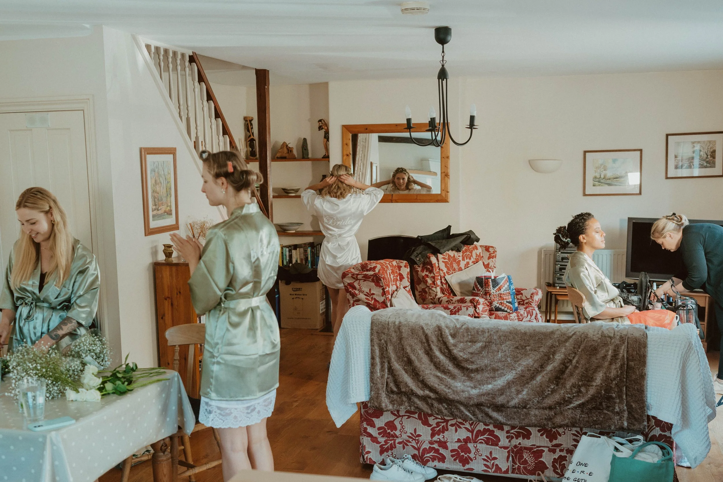 Bridal party preparing in a living room, with women in satin robes, some doing makeup and hair, and a table with flowers and a person arranging items.