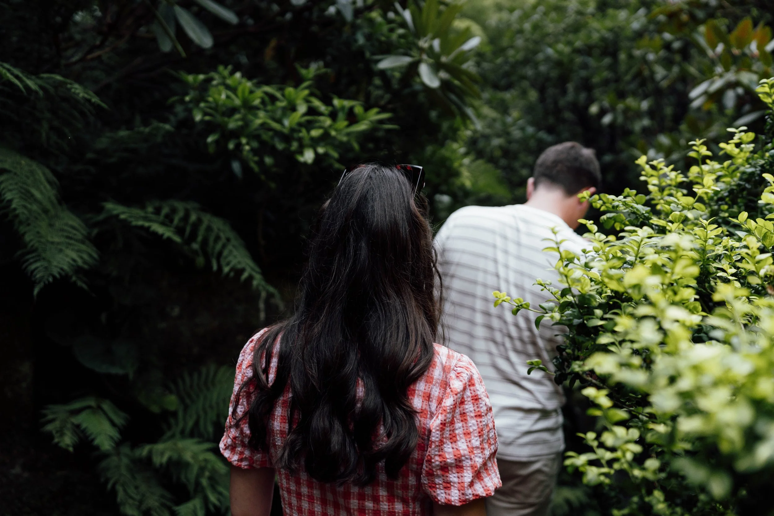 A woman with long dark hair wearing a red and white checked shirt walking in a lush green garden with a man in a white striped shirt ahead of her.