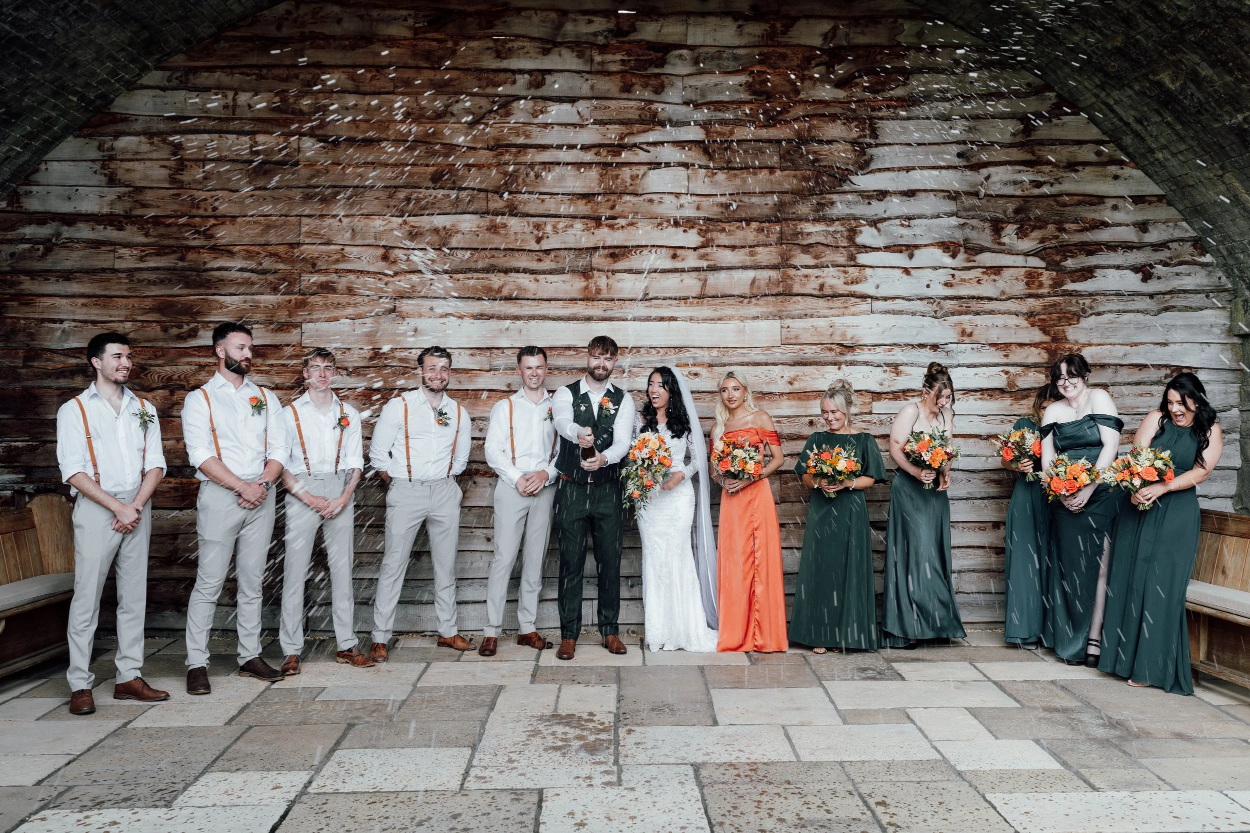A wedding party of eleven people stands together in an indoor rustic setting with a wooden wall backdrop. The bride and groom are in the center, smiling and holding a bottle of champagne, with guests and bridesmaids on either side, all dressed in for