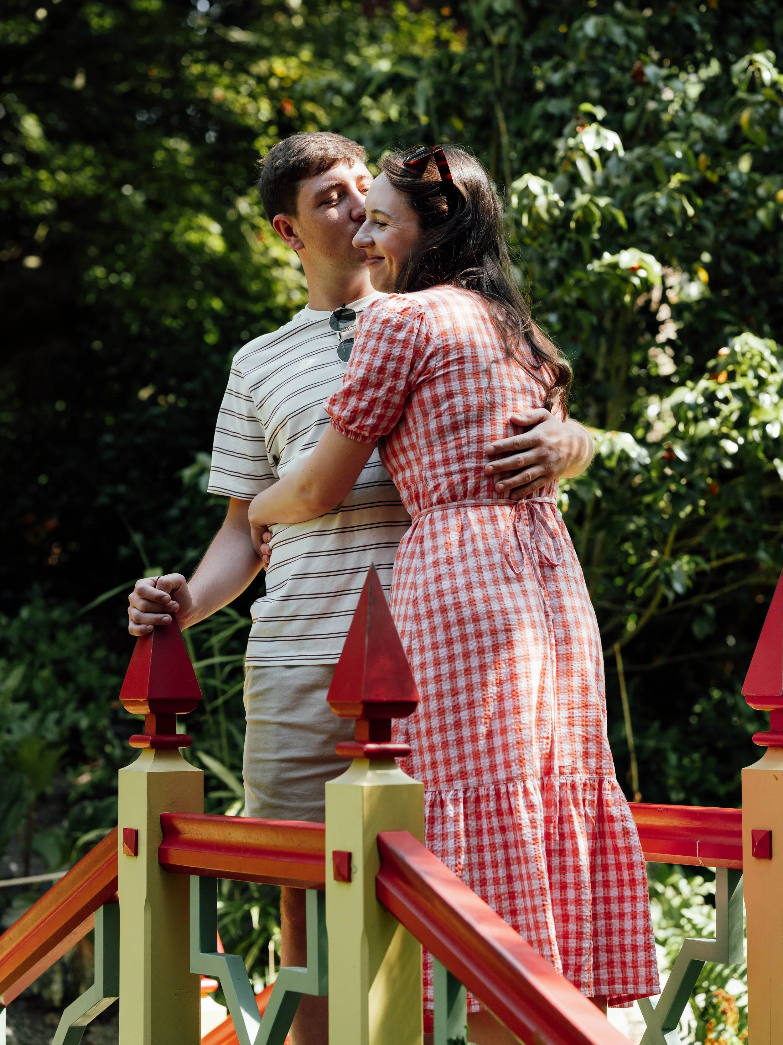 A couple standing on a small bridge in a lush green garden, embracing and sharing a kiss, with sunlight filtering through the trees.