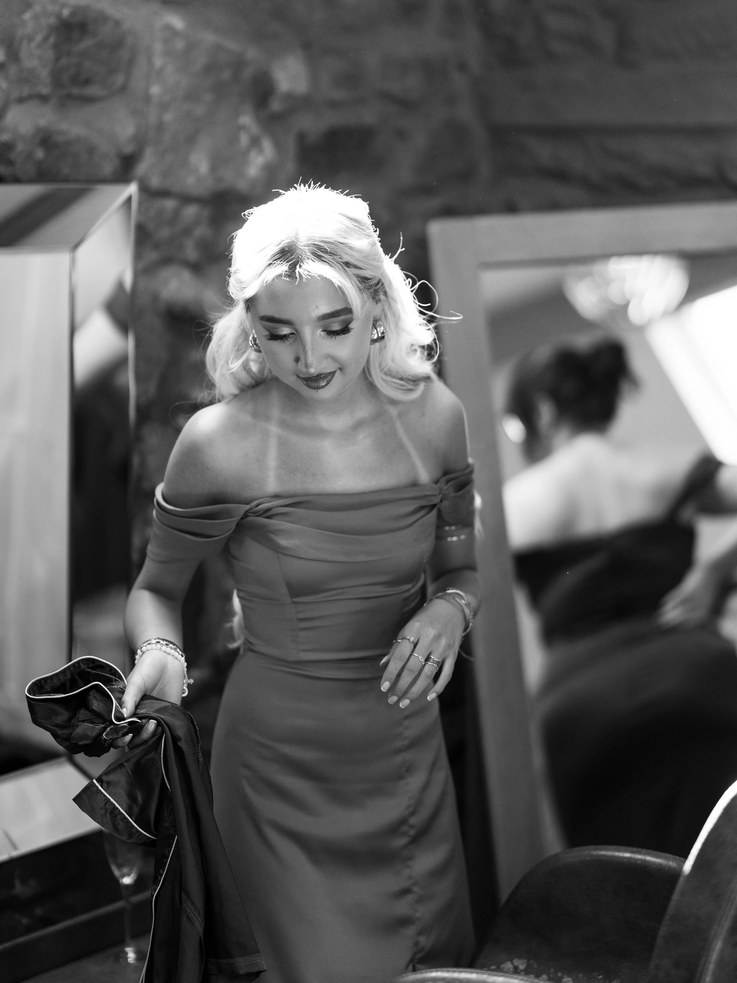 A woman with blonde hair dressed in an off-the-shoulder gown, holding a jacket, looking down, with a mirror and a rustic stone wall in the background.