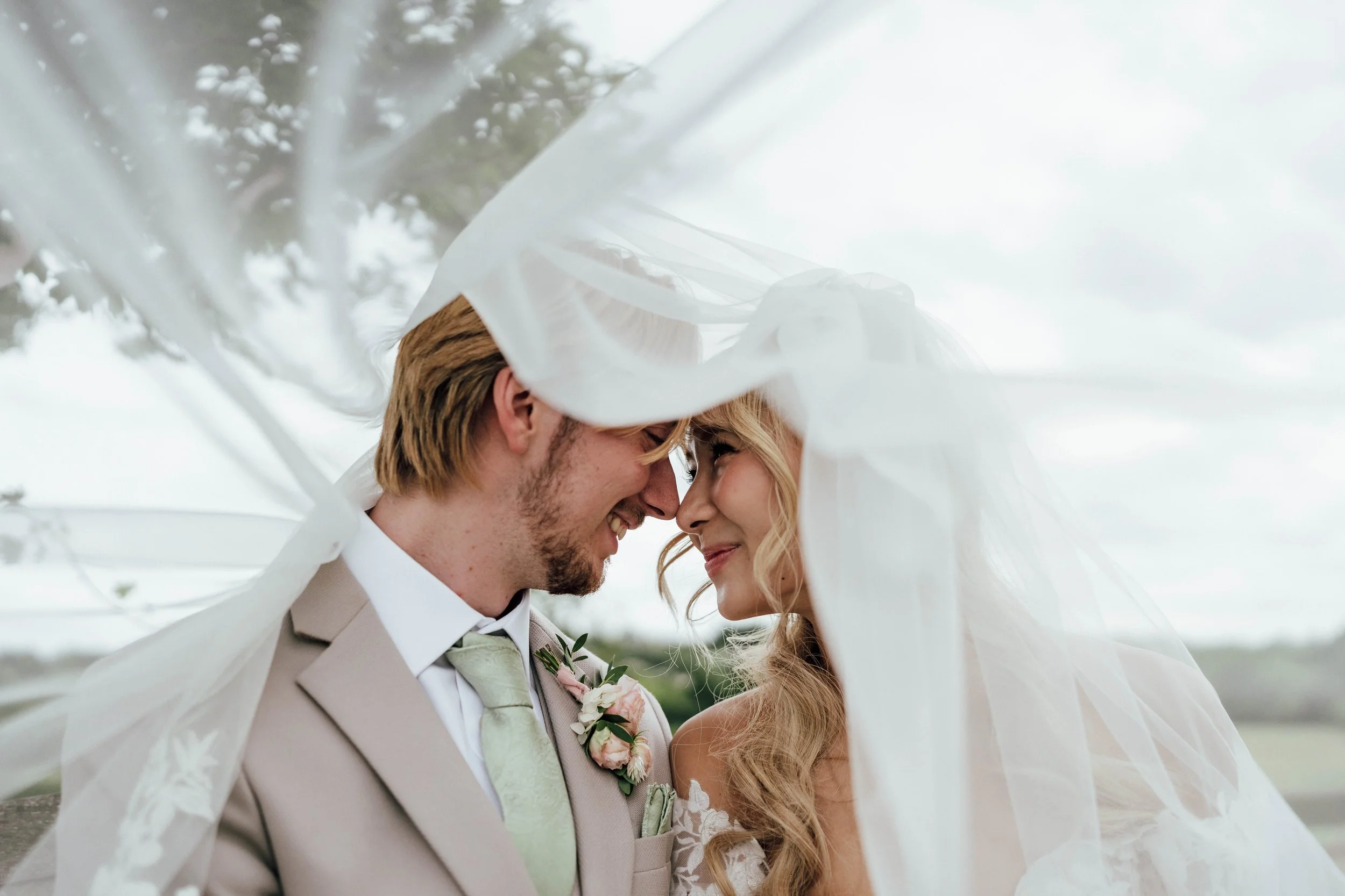 A bride and groom share a close moment under a sheer white veil outdoors during daytime.