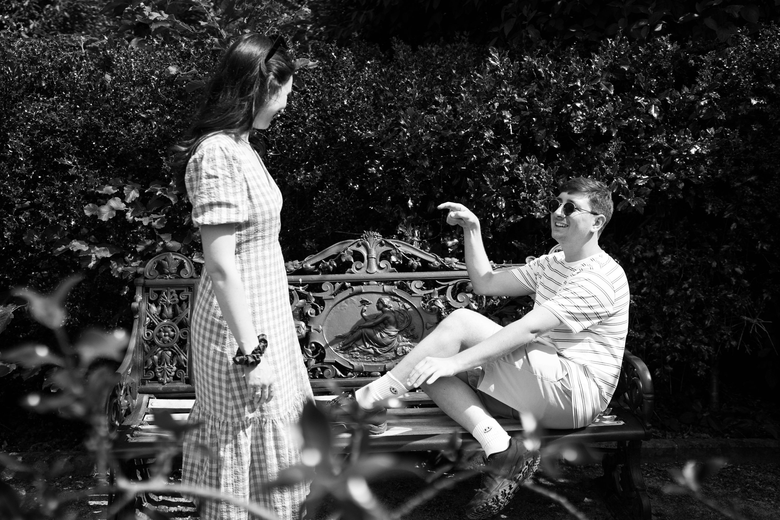 A young woman and a young man are sitting on a decorative outdoor bench with a lion motif, surrounded by bushes and trees. The woman is standing in front of the bench, smiling, while the man, wearing sunglasses, is sitting with his leg outstretched, 