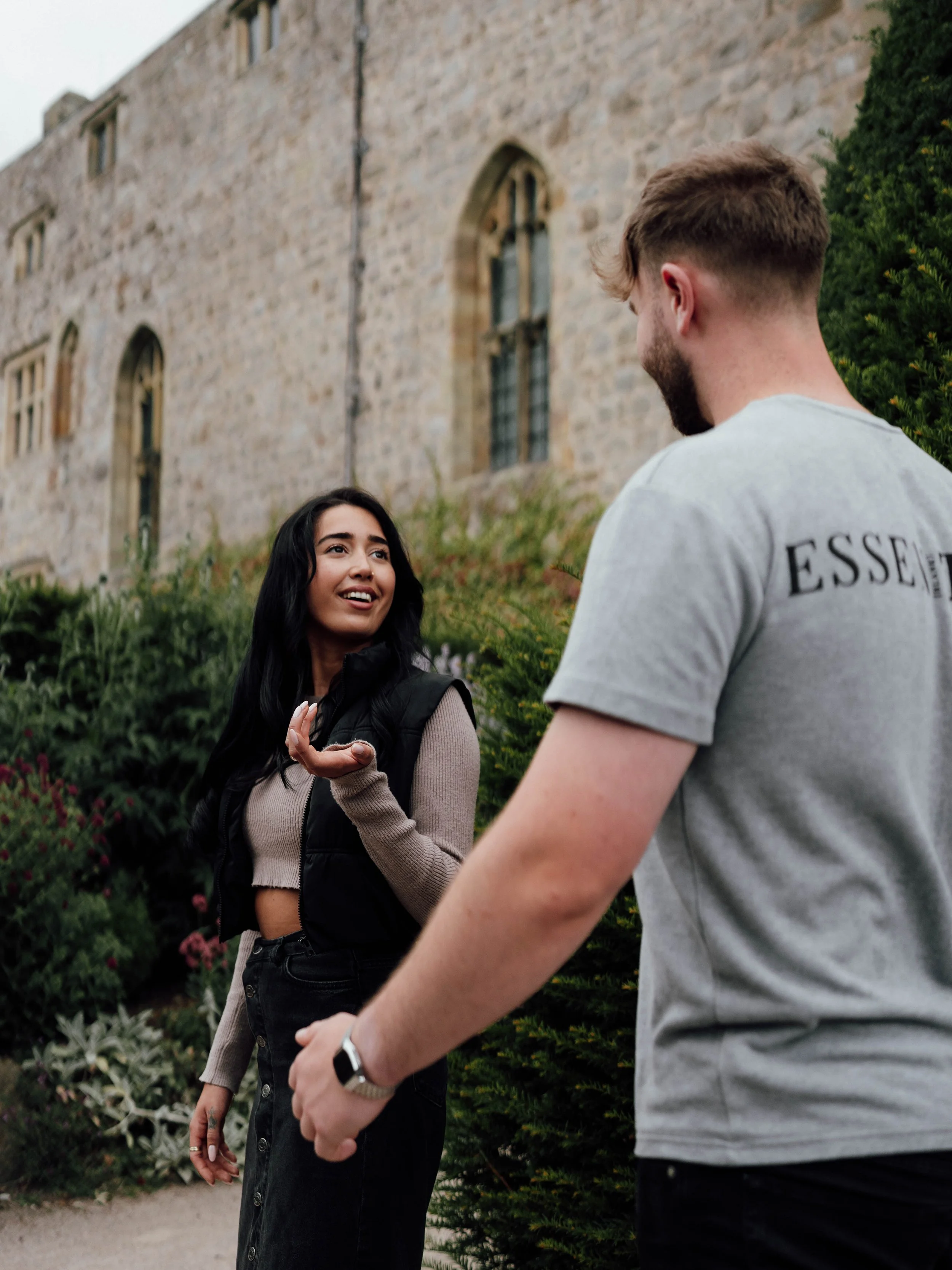A woman with long dark hair, wearing a beige cropped sweater, black skirt, and black vest, is talking to a man with short brown hair, wearing a gray t-shirt and a smartwatch, outside near a stone building and green bushes.