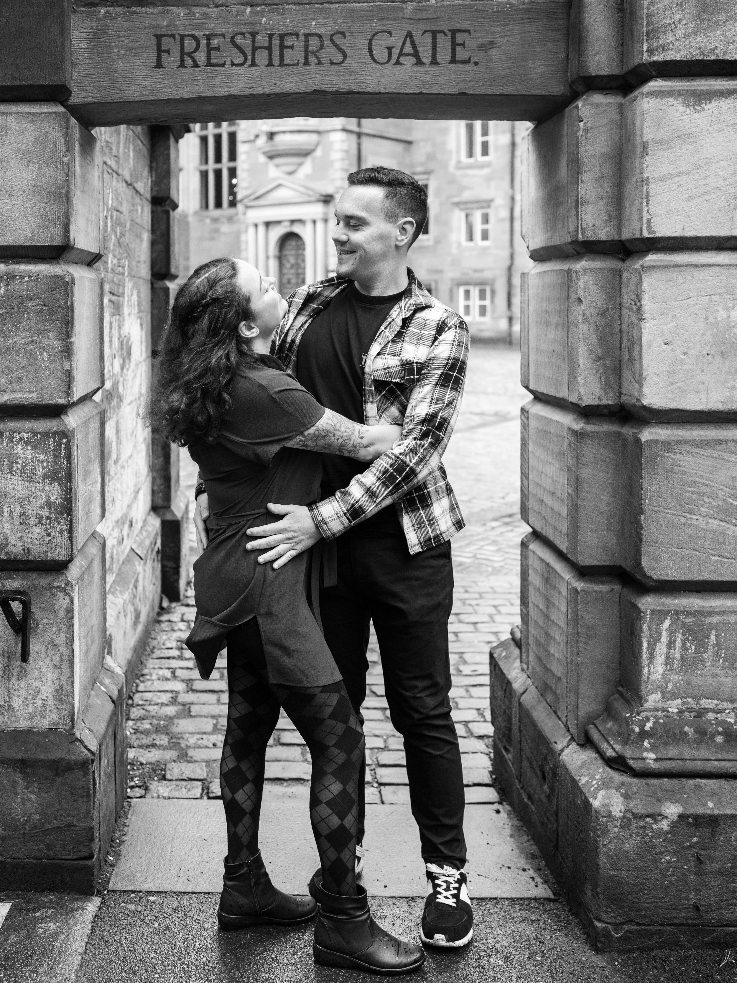 A couple sharing a joyful moment under a stone archway labeled "Freshers Gate" in black and white.