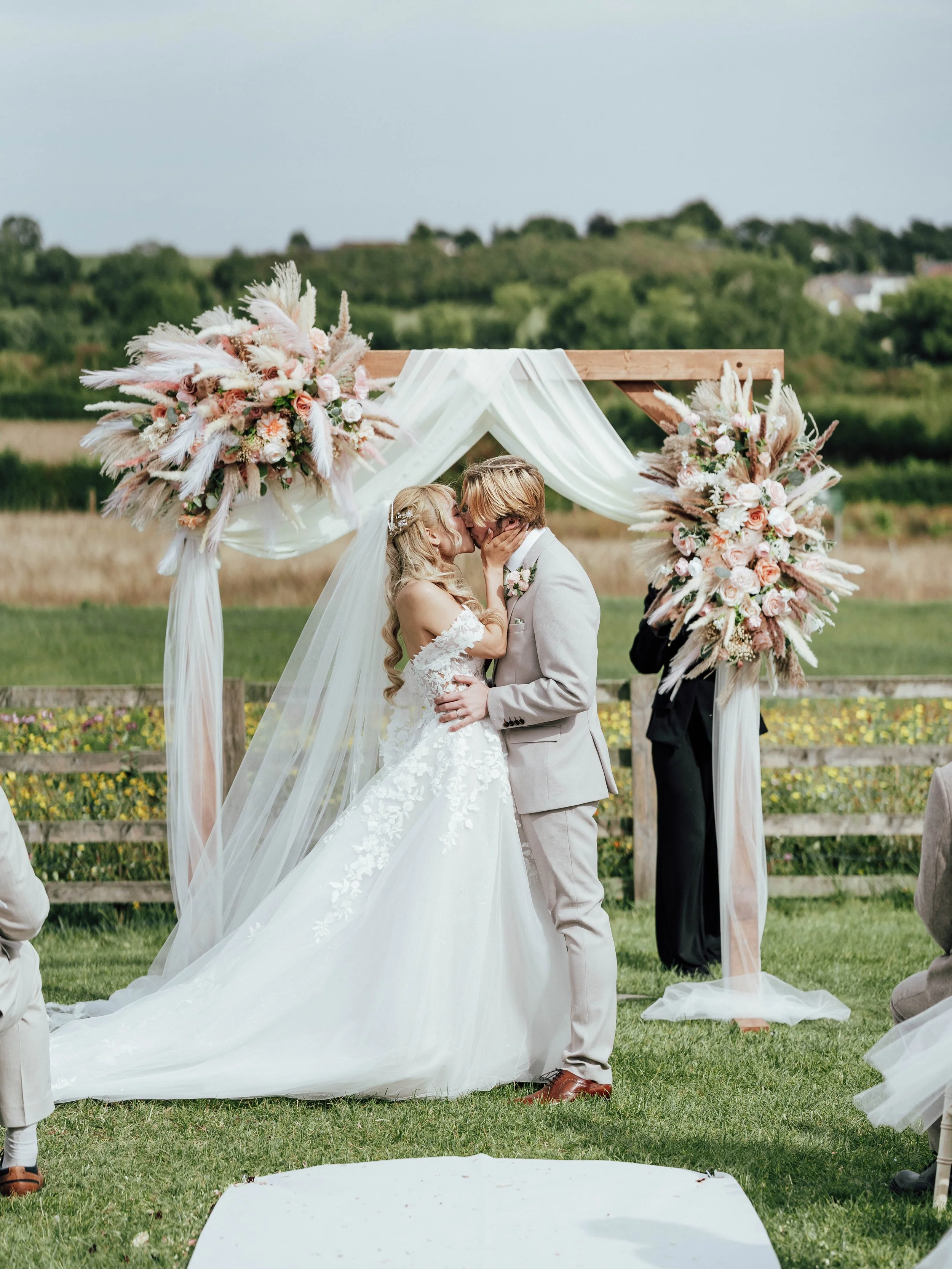 A bride and groom kiss during an outdoor wedding ceremony under a wooden arch decorated with pink and white floral arrangements and draped white fabric.