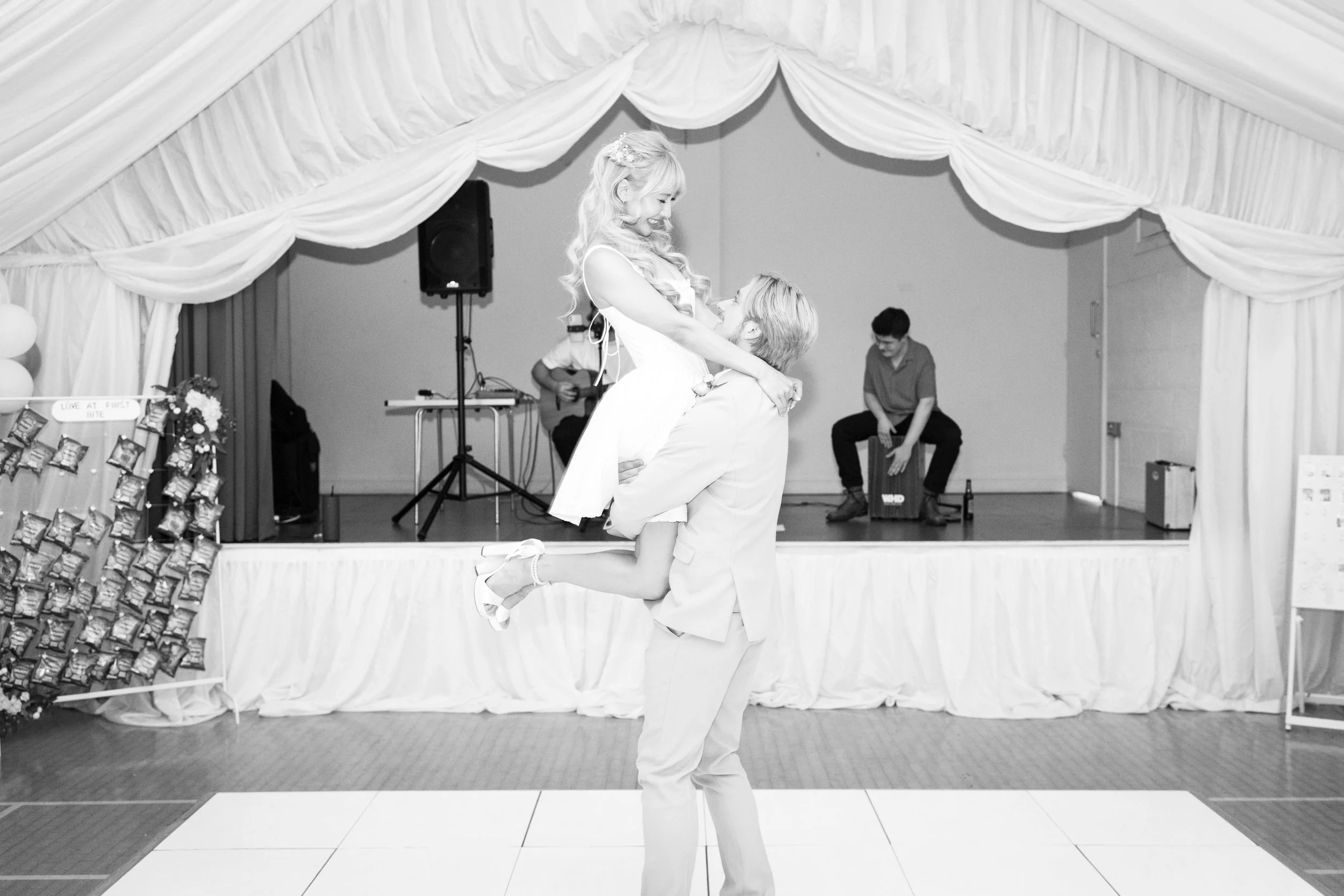 A man lifts a woman in a white dress during a celebration. She has long, curled hair. The background features a stage with musicians and a string of balloons.