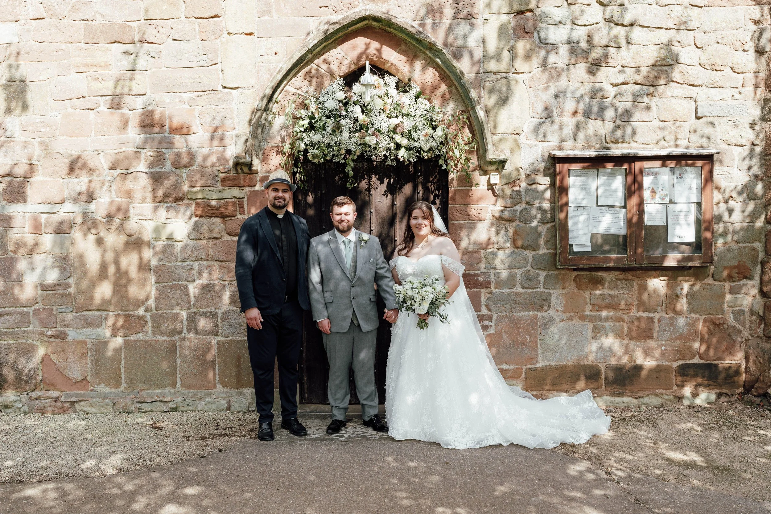 A wedding scene featuring three people in front of a stone wall with arched doorway decorated with white flowers. The bride in a white wedding gown holding a bouquet, a groom in a light gray suit with a white shirt, and a man in a black suit with a g