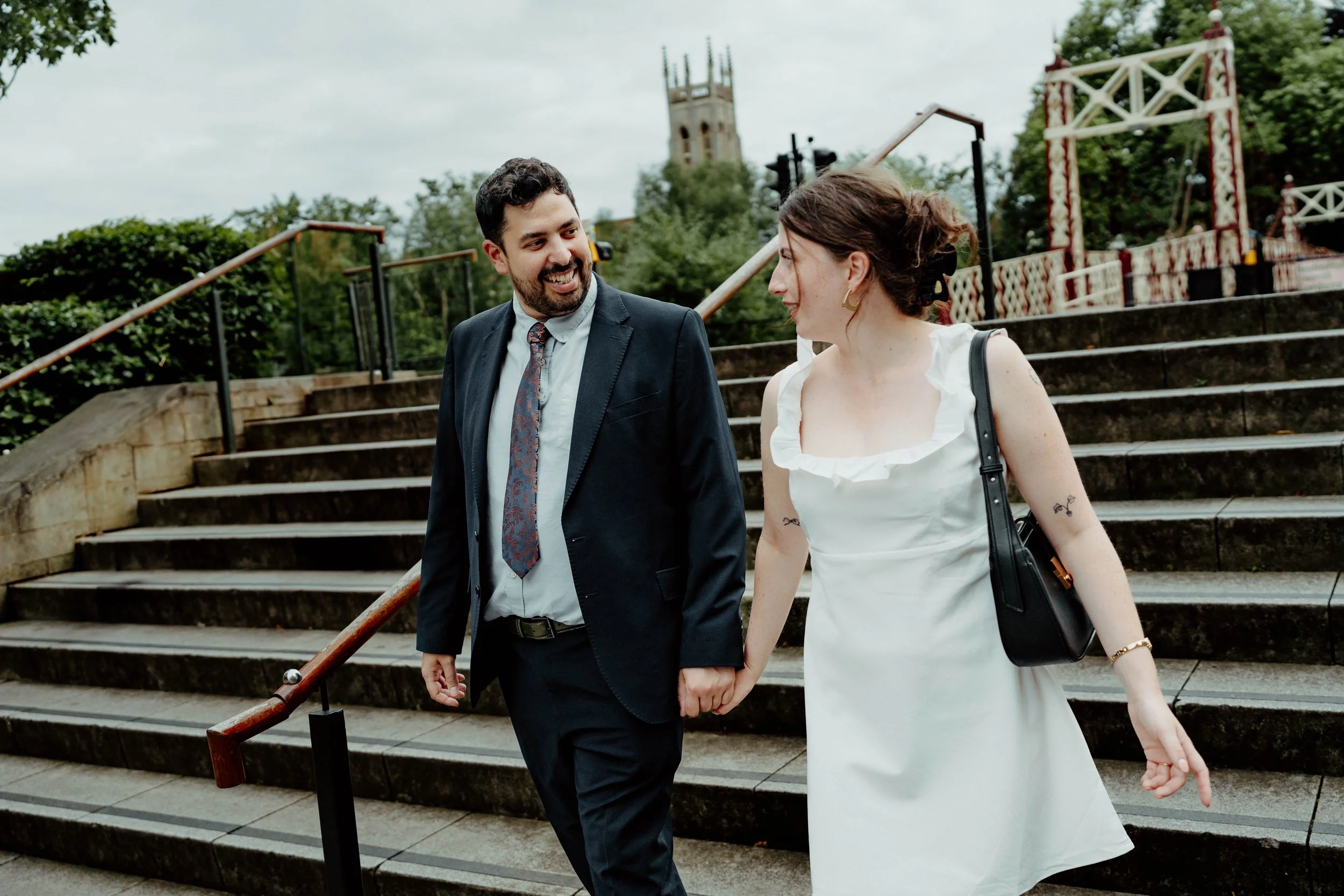 A couple holding hands and walking down outdoor stairs, smiling and looking at each other on a cloudy day.