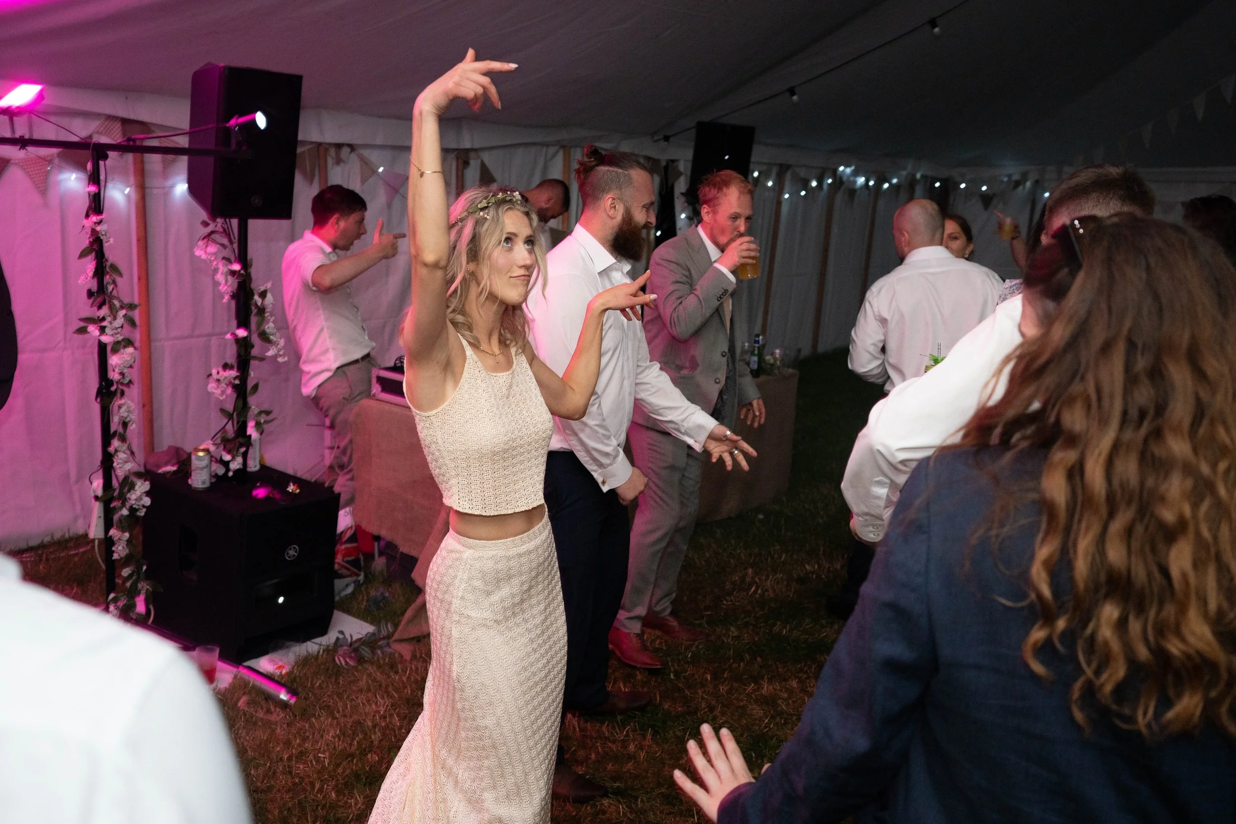 People dancing at a party under a tent with string lights, a woman in a cream dress is front and center with her arm raised, surrounded by others enjoying the music.