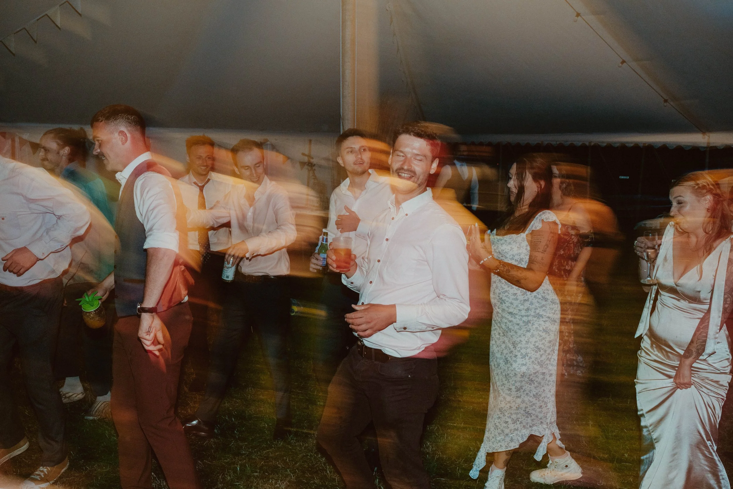 People dancing and socializing at a party under a large tent, with some holding drinks and smiling.