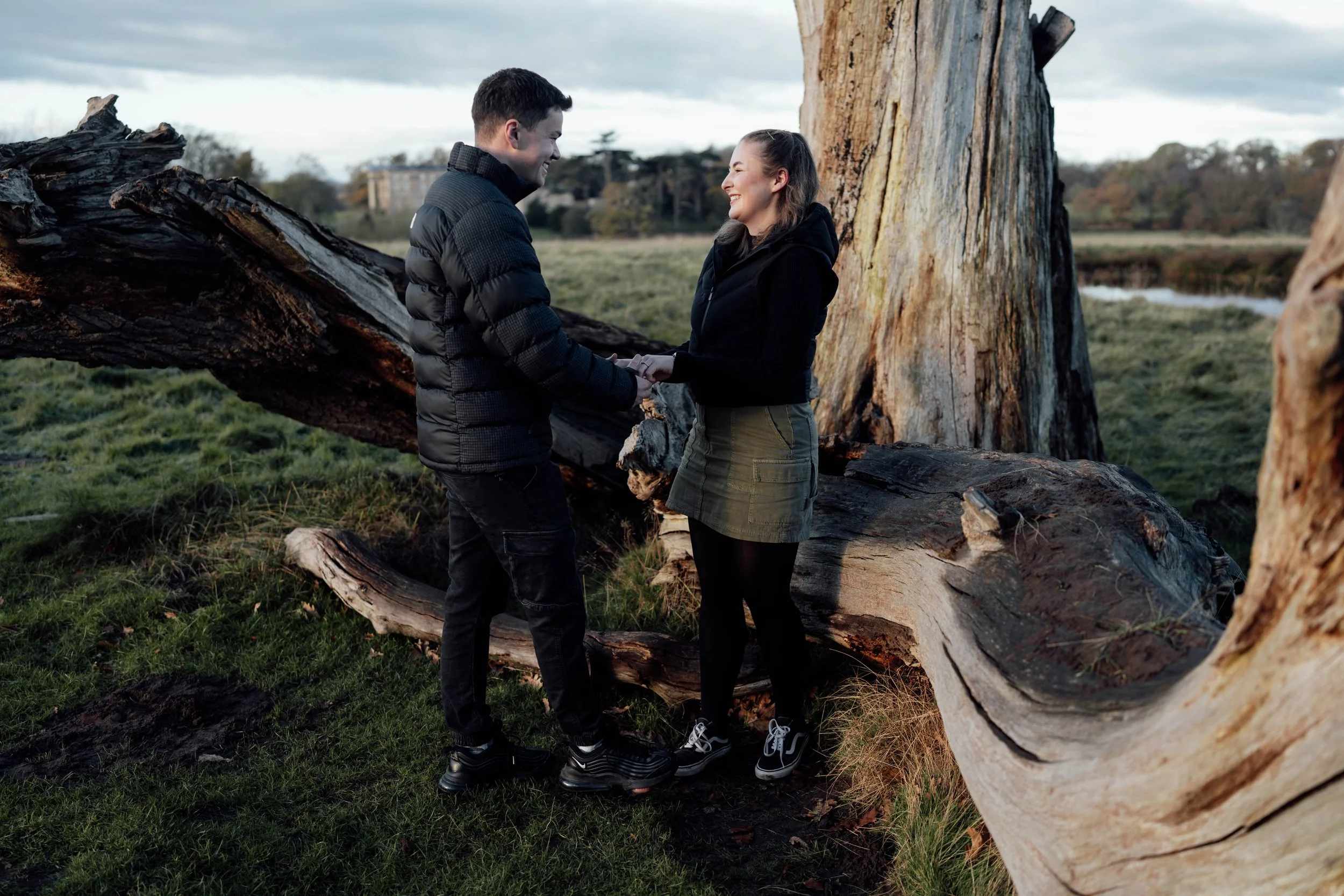 A couple holding hands and smiling at each other outdoors near a fallen large tree trunk with a grassy field and trees in the background.