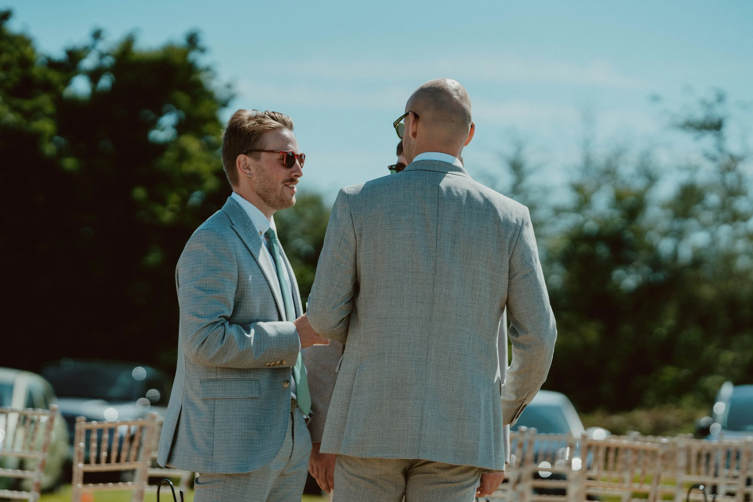 Two men in suits having a conversation outdoors near parked cars and trees on a sunny day.