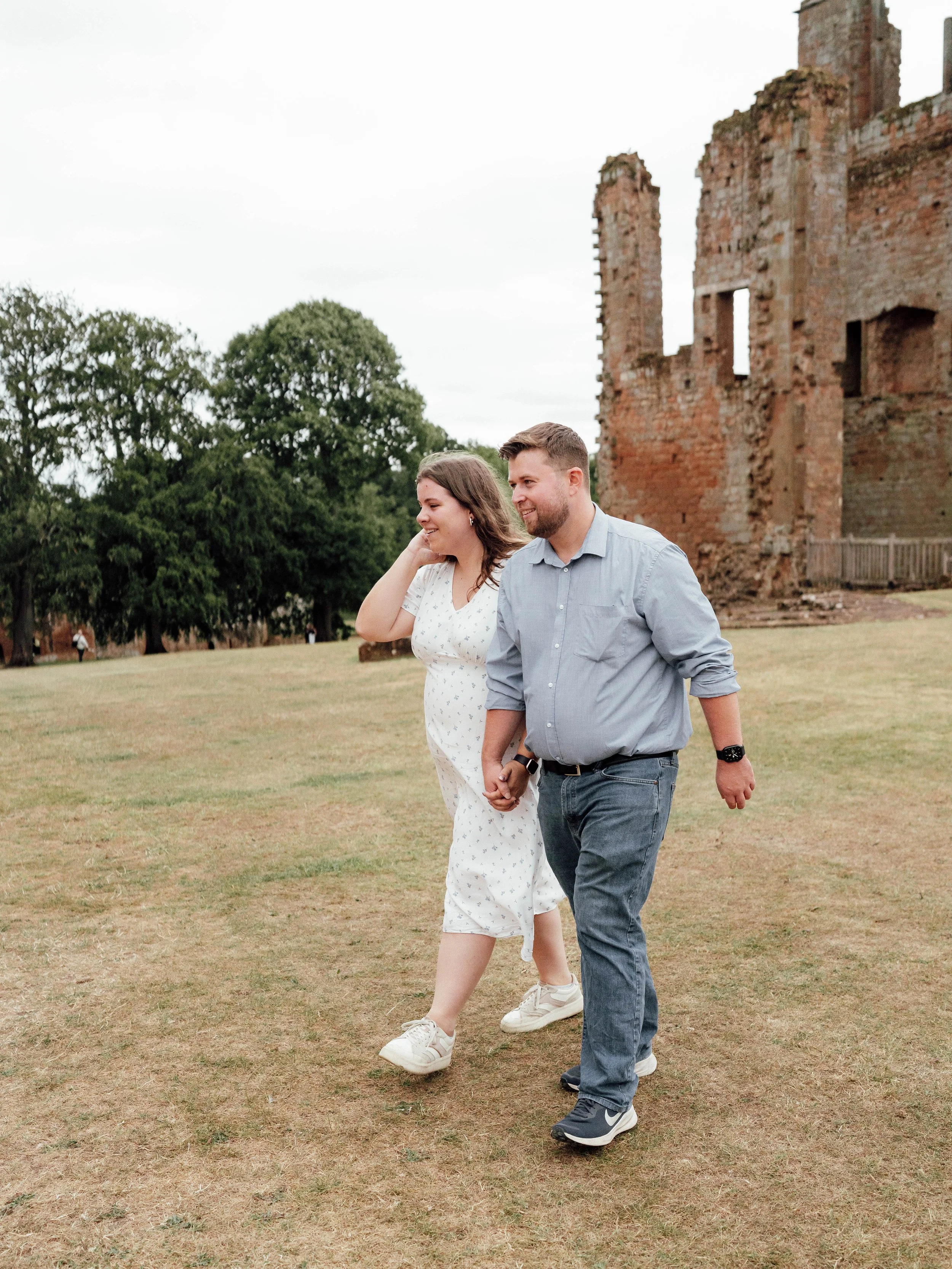 A couple holding hands and walking on grass in front of a ruined brick building with trees in the background.