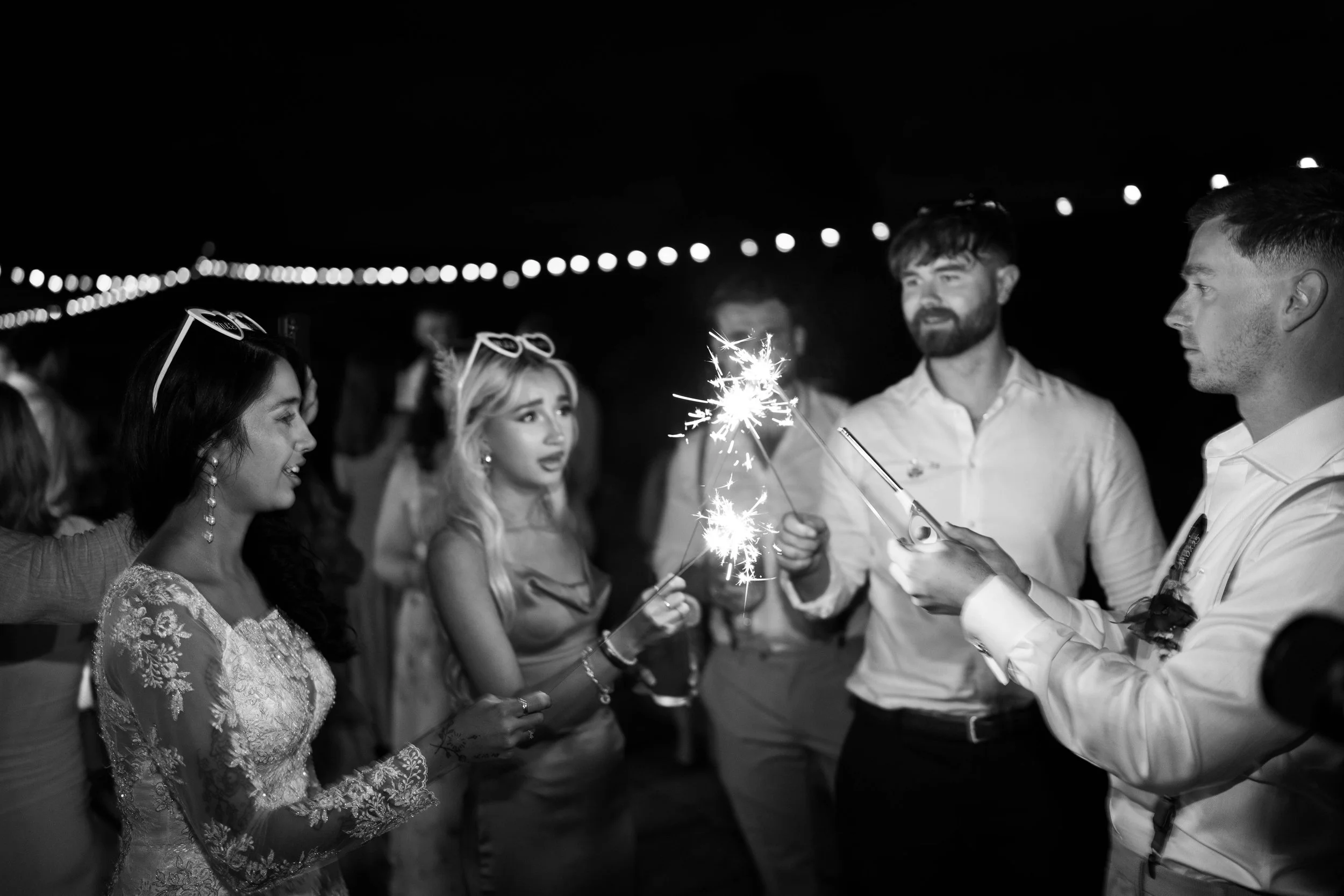 Group of people celebrating at night with sparklers, dressed in formal attire, with string lights overhead.