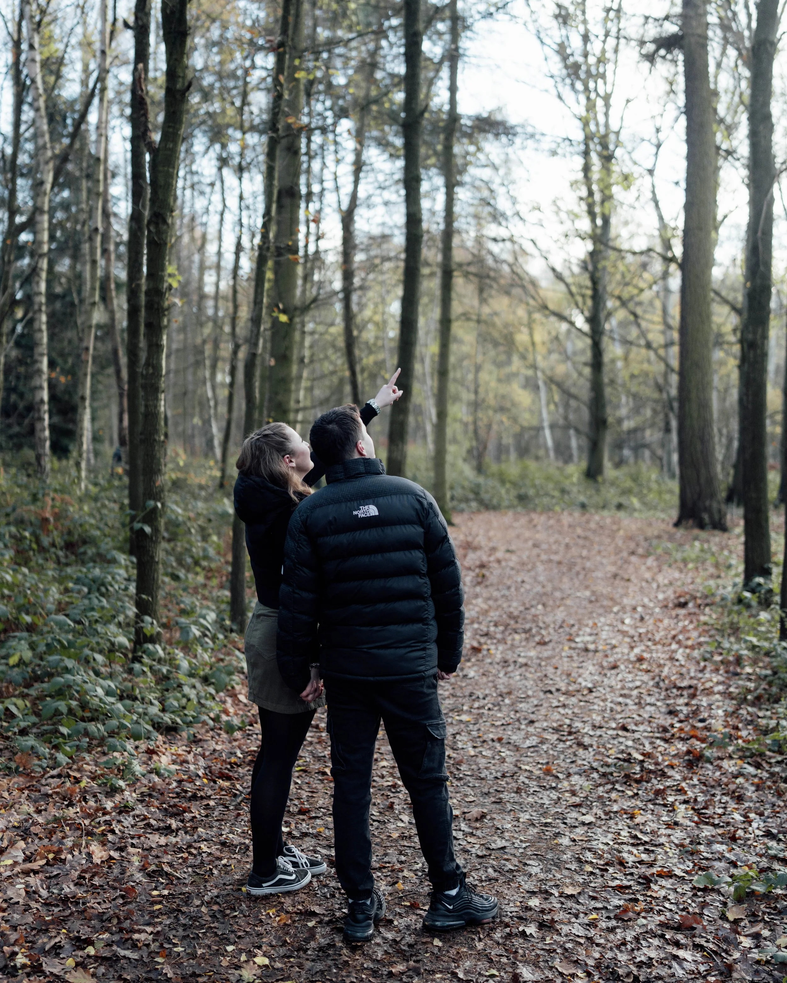 A man and woman in outdoor jackets standing on a leaf-covered forest trail, with the woman pointing at something in the tall trees.