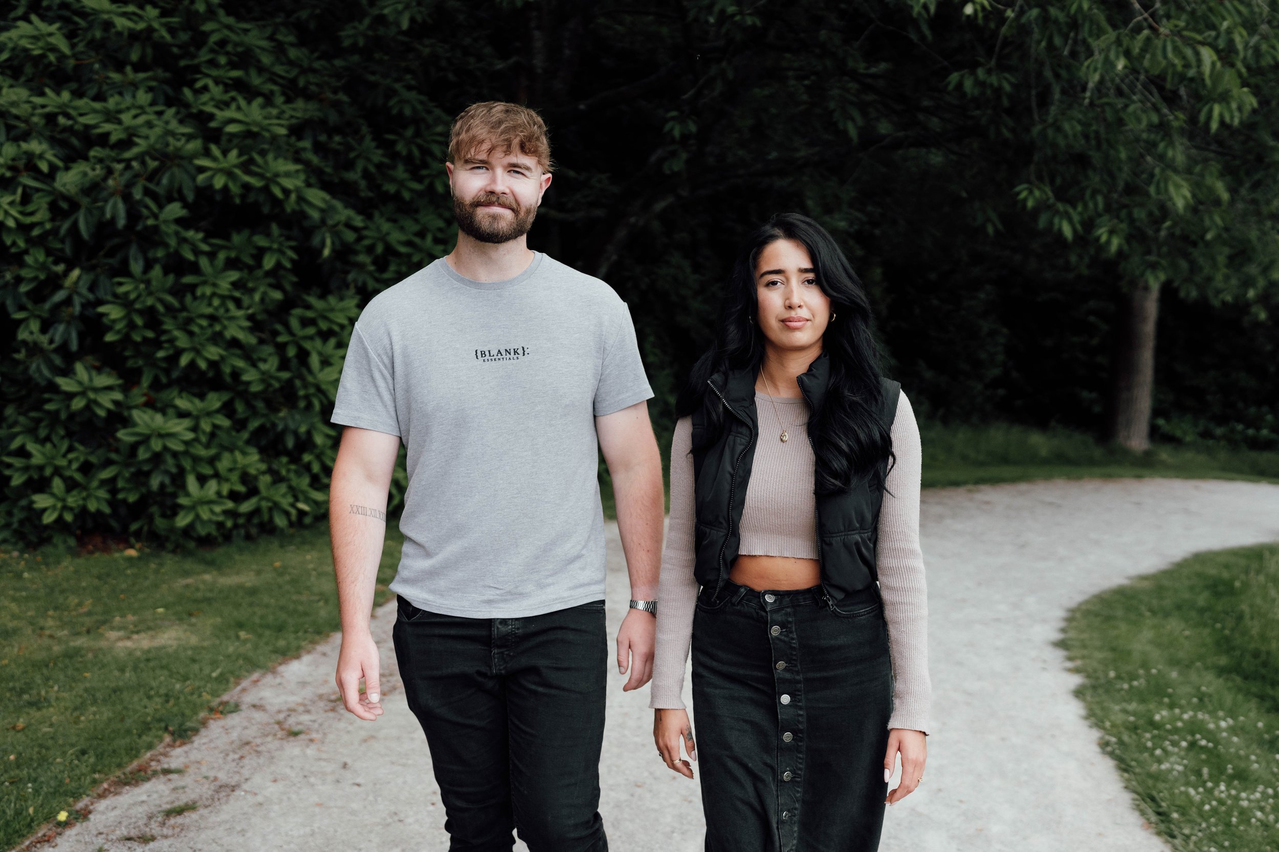 A young man and woman walking outdoors on a dirt path surrounded by green foliage. The man has curly hair and a beard, wearing a light gray t-shirt and black pants. The woman has long black hair, wearing a beige crop top, black vest, and a black skir