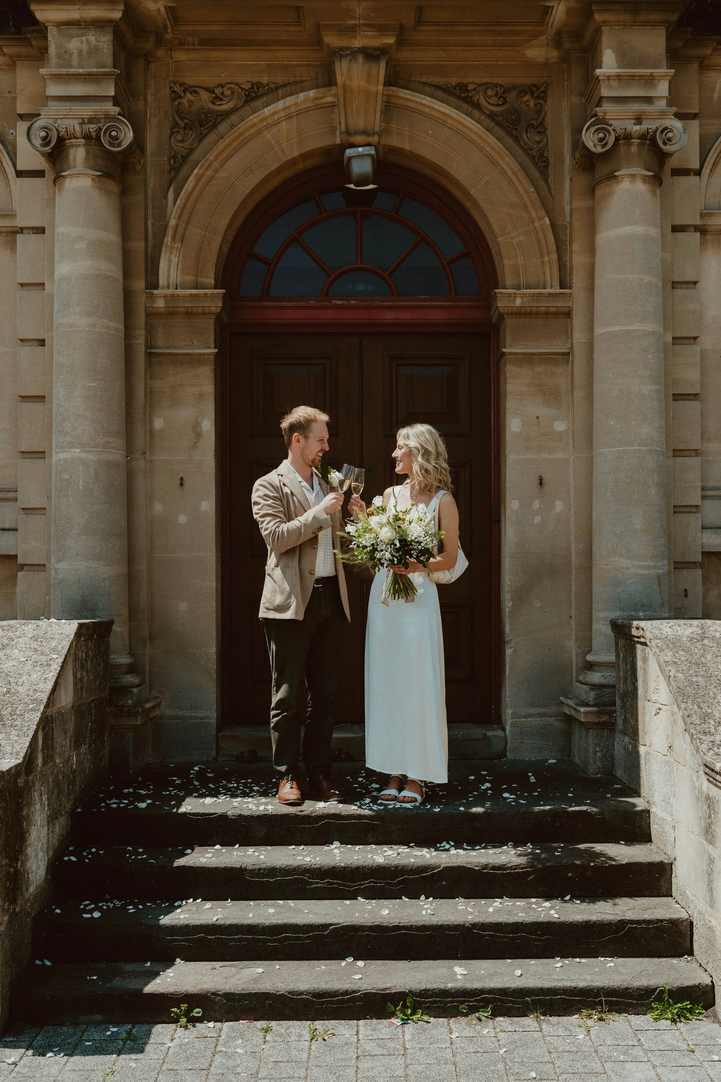 A newlywed couple celebrating on the steps of a historic building, holding wine glasses and a bouquet with white flowers, surrounded by scattered flower petals.