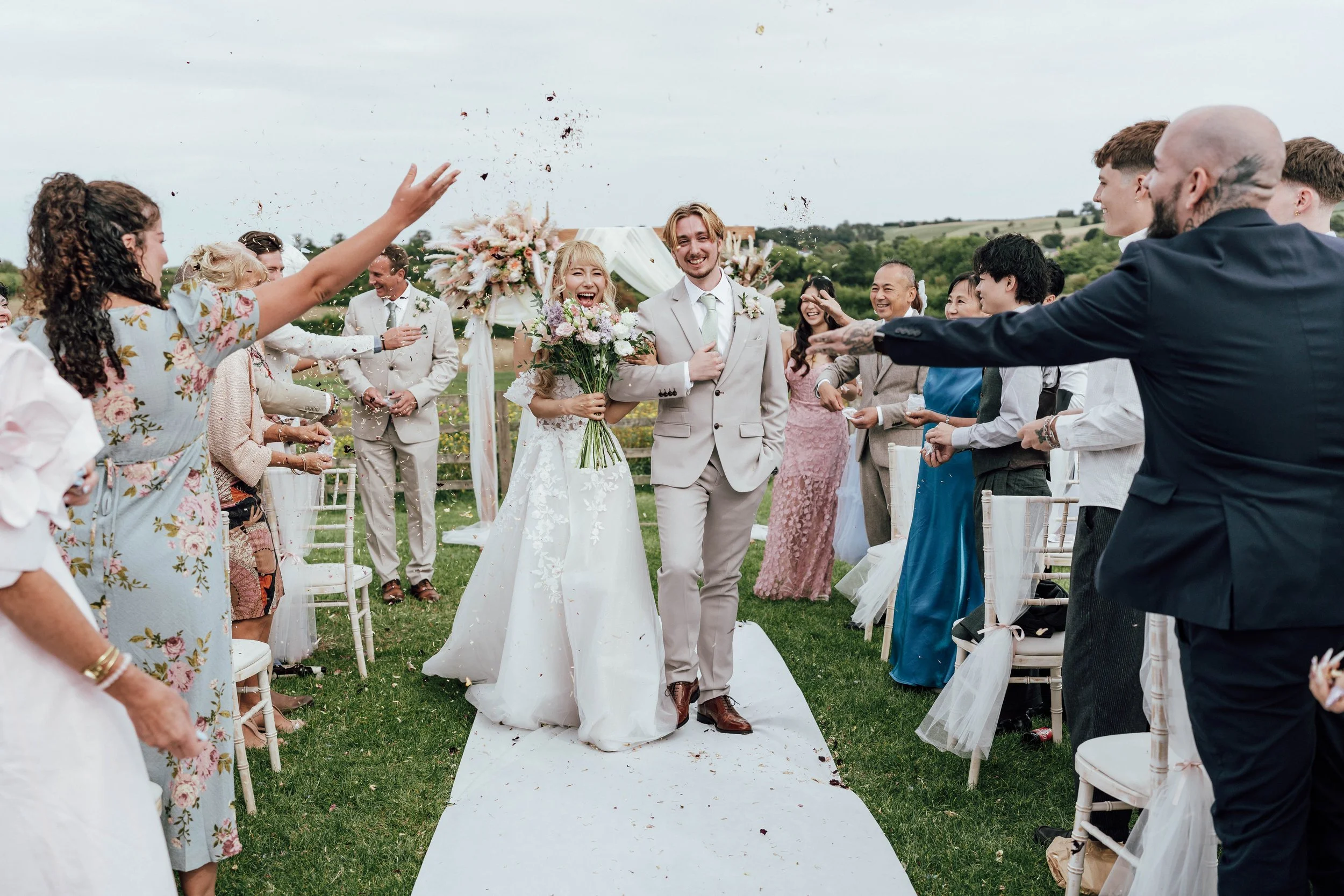 A newlywed couple walking down the aisle together outside, surrounded by friends and family throwing flower petals. The bride is holding a bouquet of flowers, and they are both smiling.