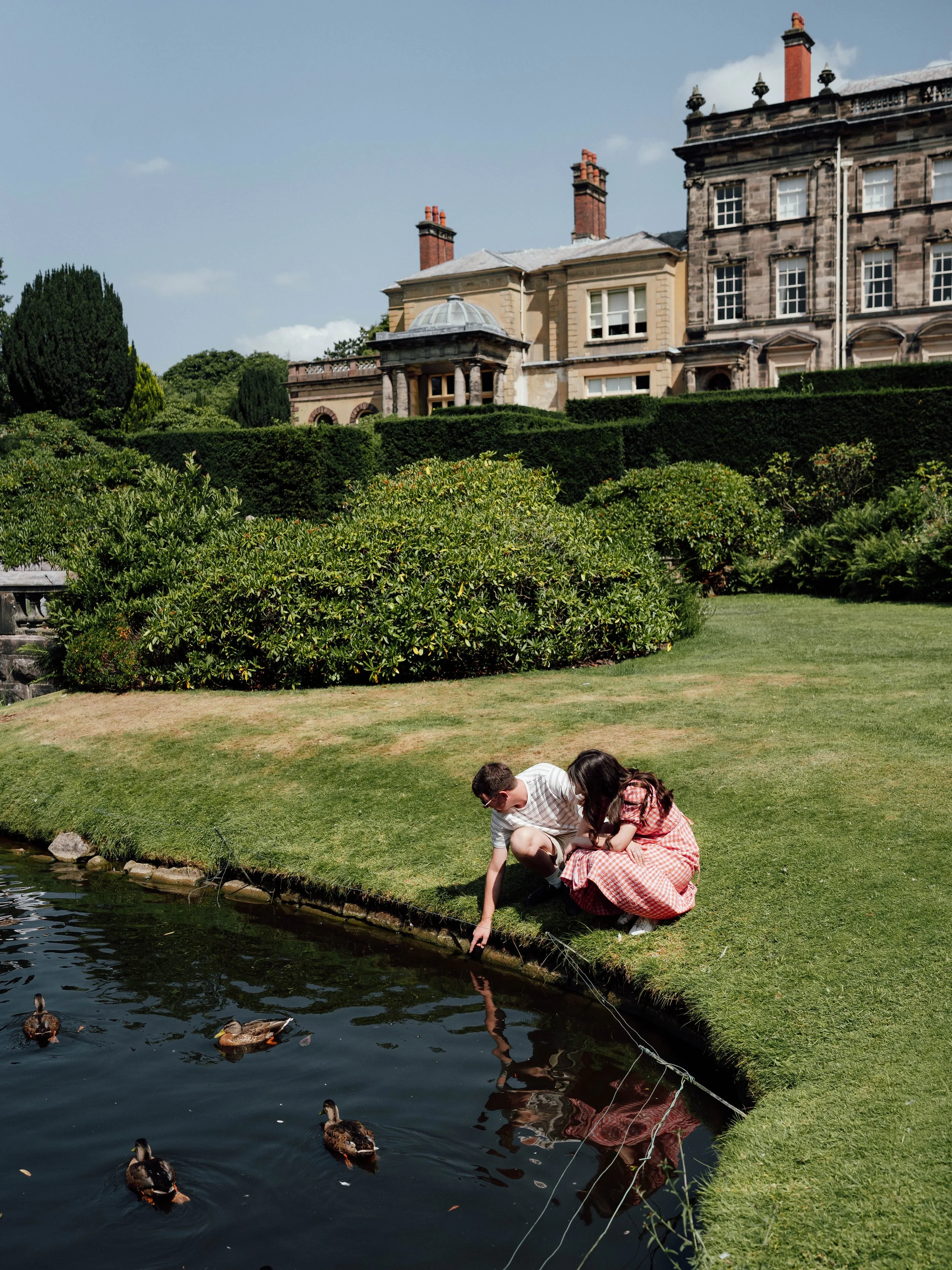 Two children, a boy and a girl, kneeling by a pond and feeding ducks in a lush green garden in front of a large historic building.