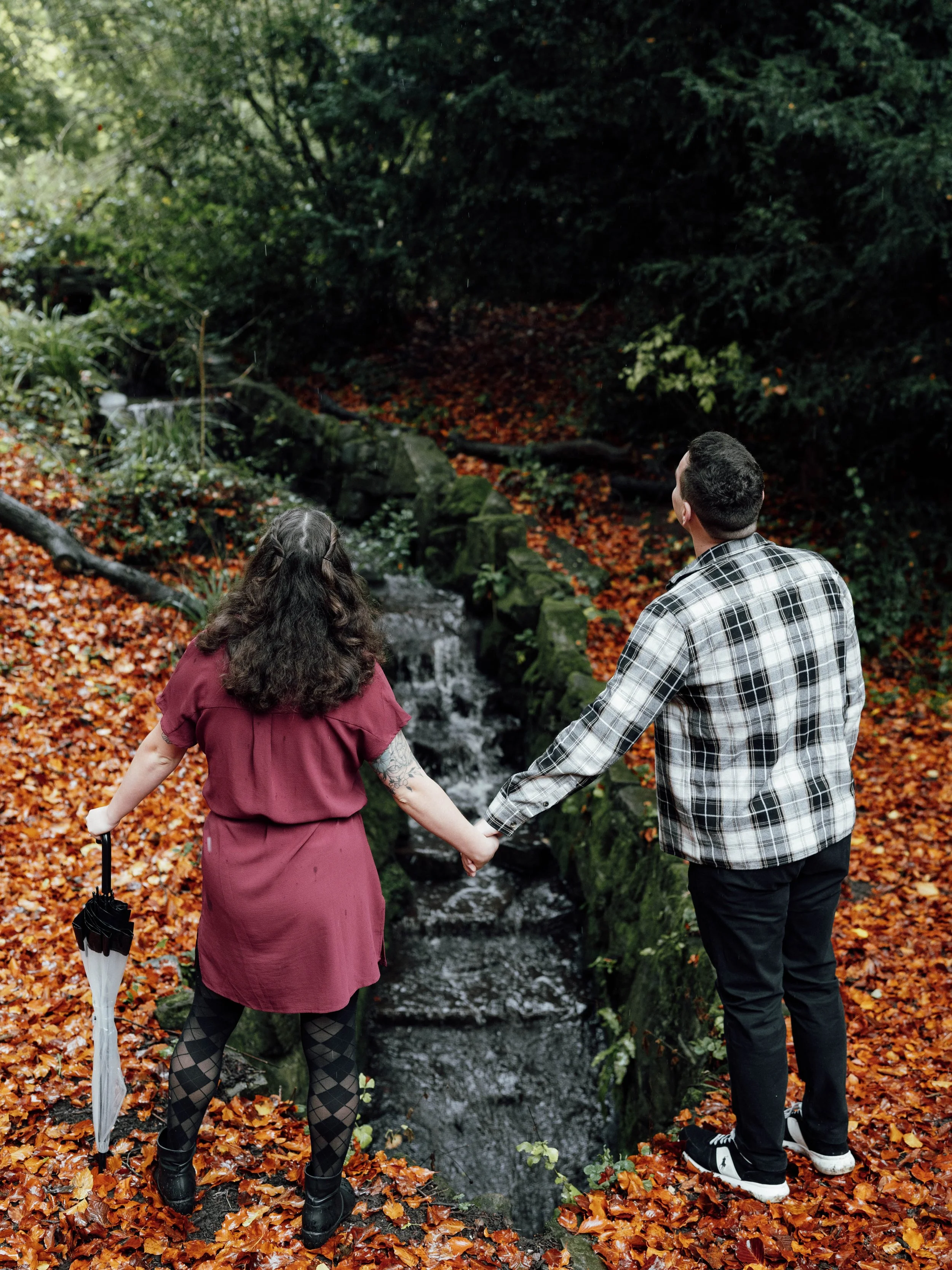A woman and a man holding hands by a small waterfall in a forest during autumn, with colorful fallen leaves on the ground.