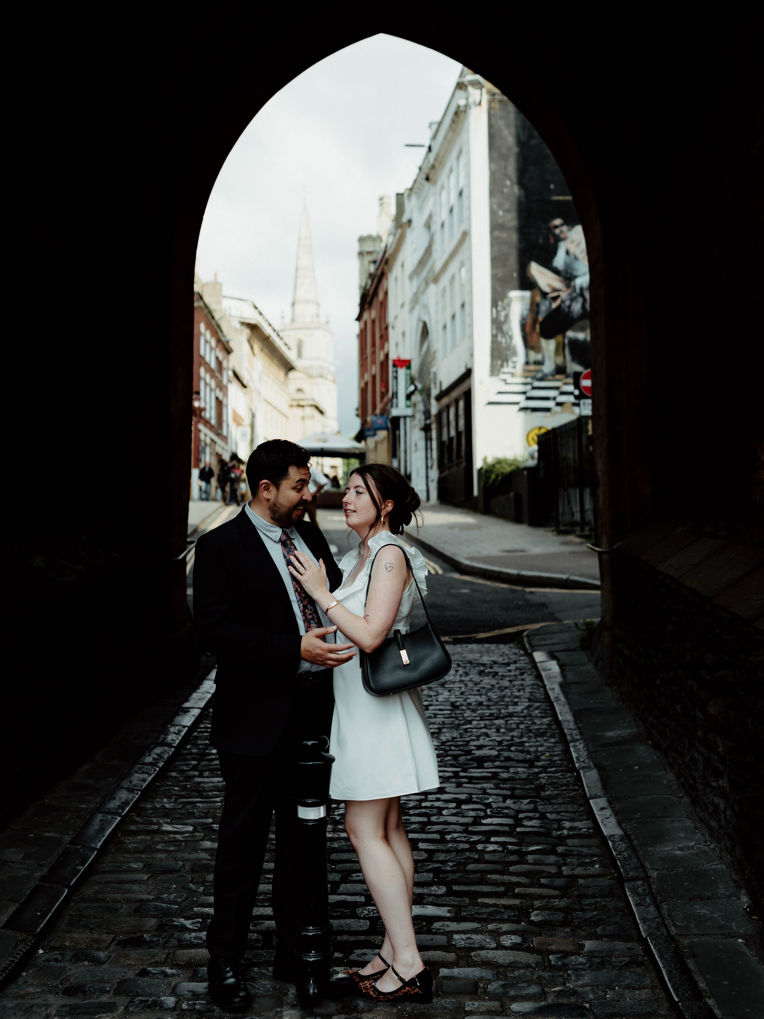 A couple is standing close together in a cobblestone street under a dark archway, smiling at each other with buildings and a church spire in the background.