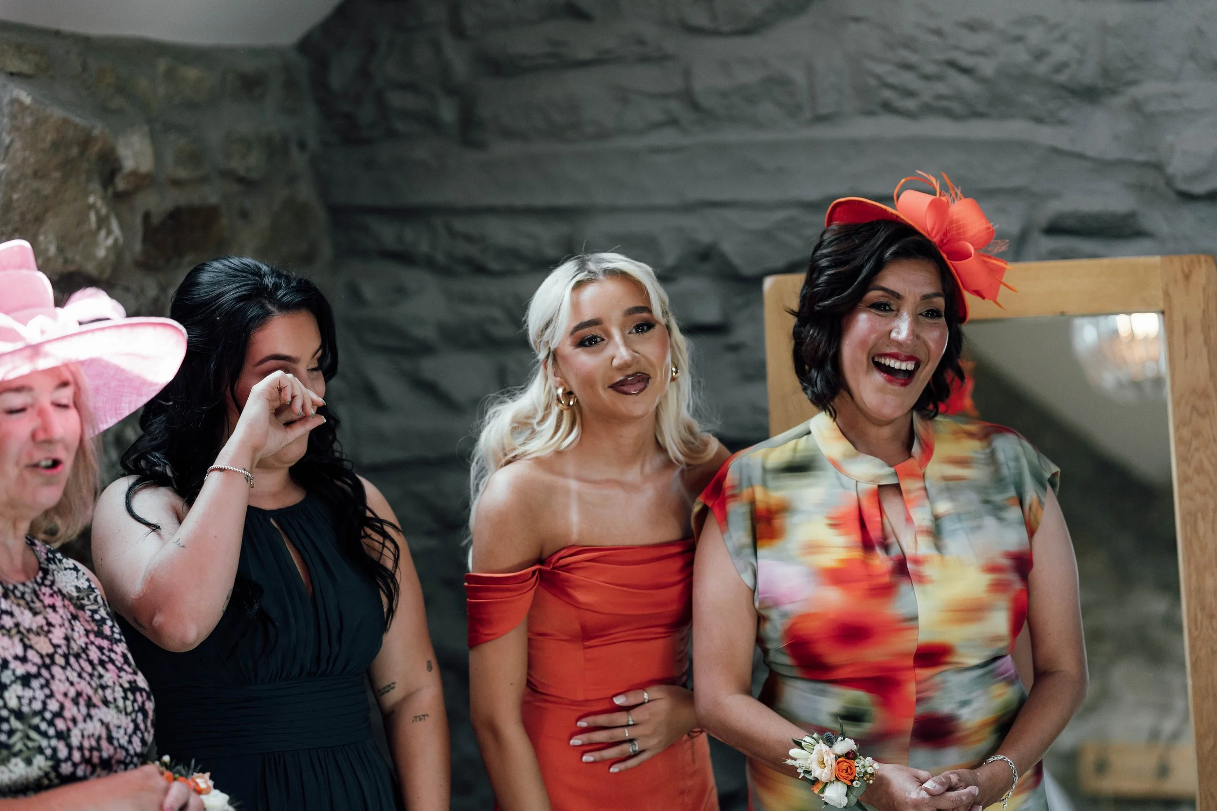 Group of women at a celebration, smiling and dressed in colorful attire, with some wearing floral accessories and a pink hat.