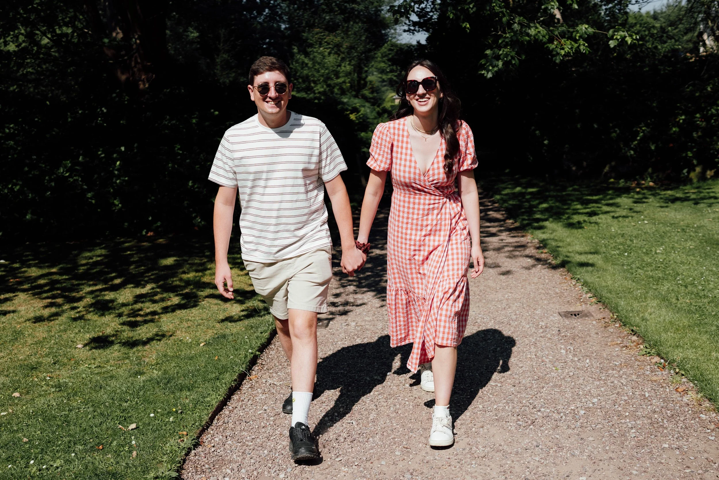 A young man and woman holding hands and walking on a gravel path in a park, surrounded by green trees and grass, smiling and enjoying a sunny day.