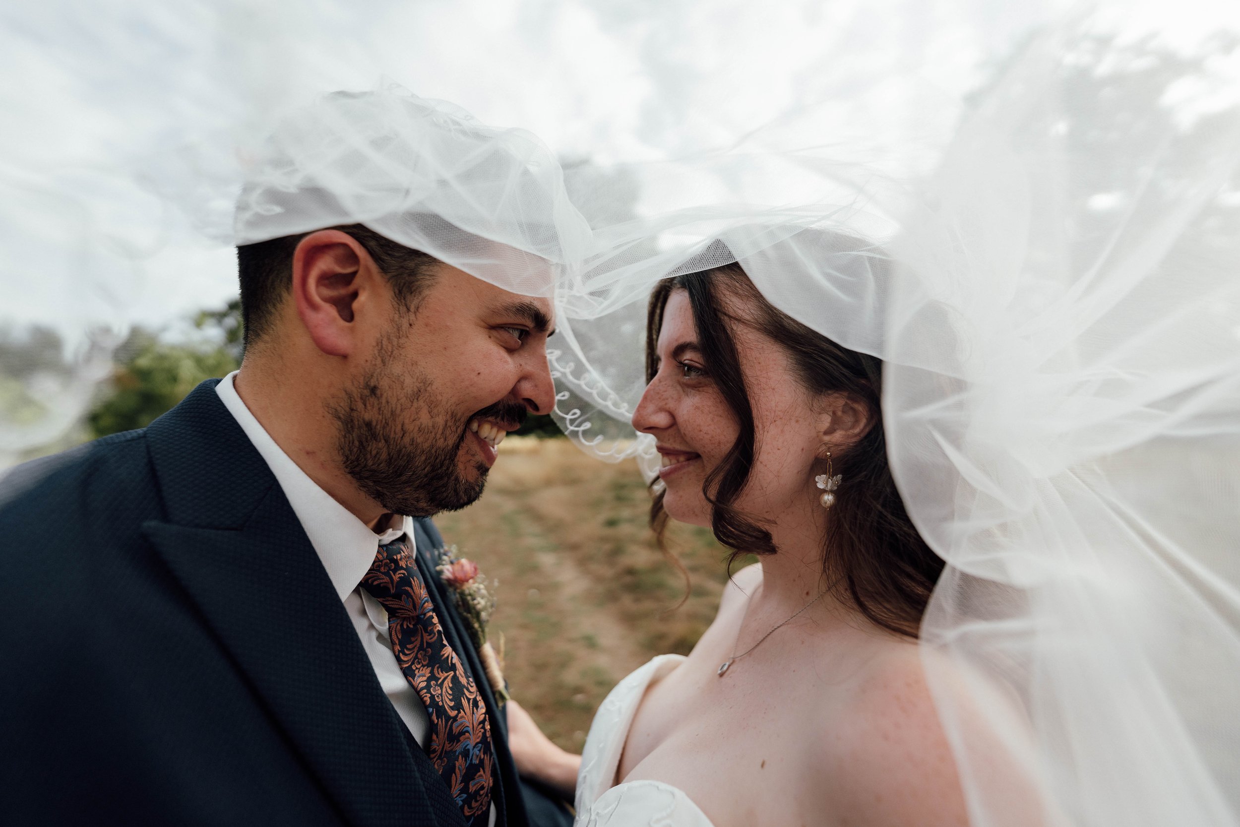 A bride and groom smiling close together under a white veil outdoors.