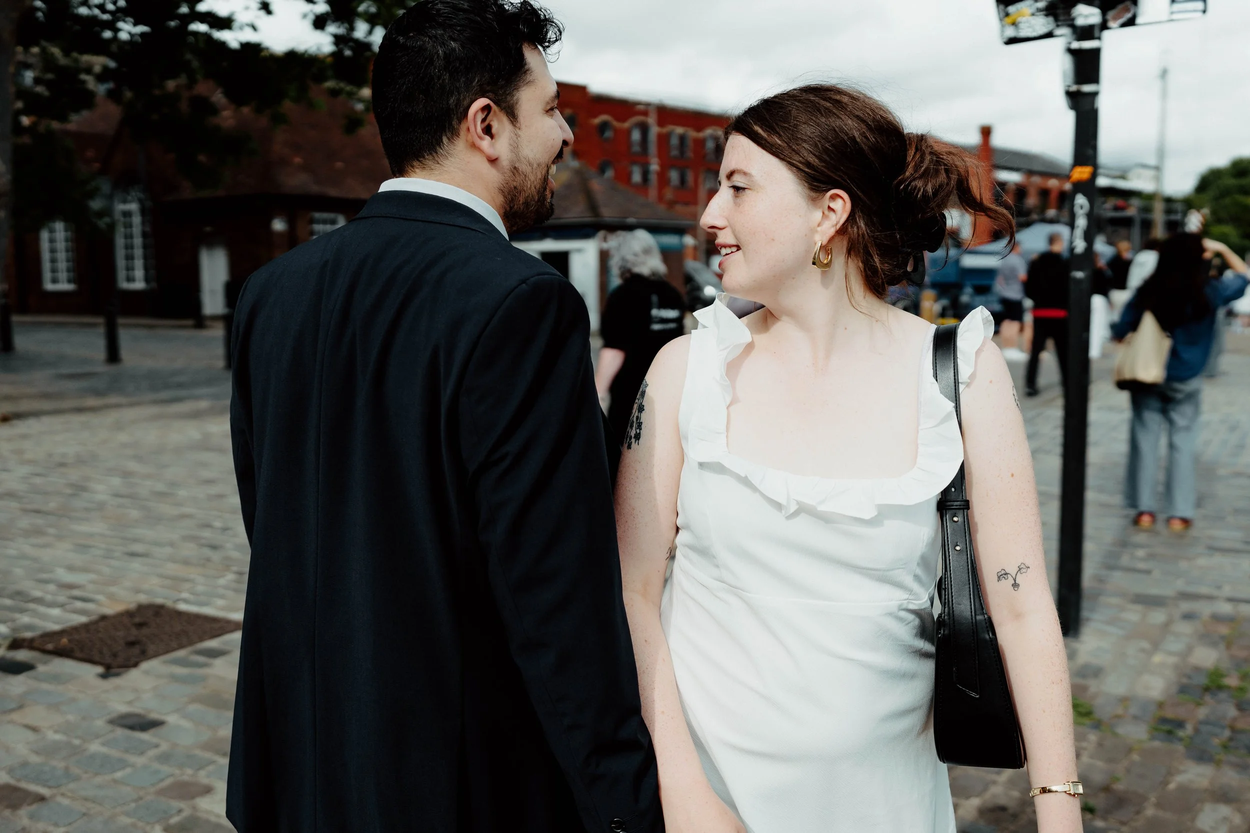 A couple standing close and looking at each other, smiling. The man is dressed in a dark suit, and the woman is wearing a white dress with ruffled edges and a black shoulder bag. They are on a cobblestone street with other people and buildings in the
