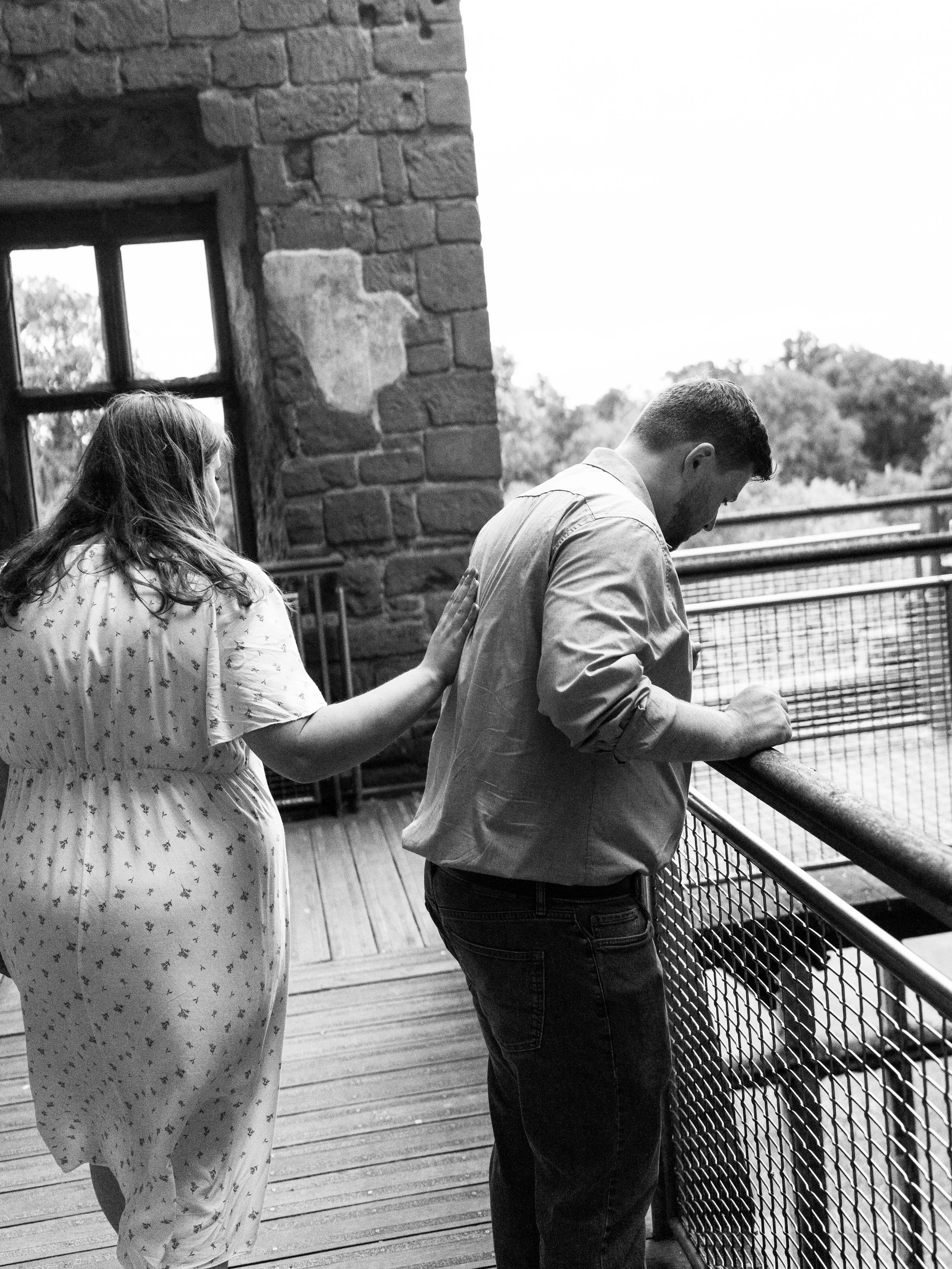 A woman places her hand on the back of a man while they look out over a railing on a balcony or observation deck in a building with brick walls, with trees visible in the background.