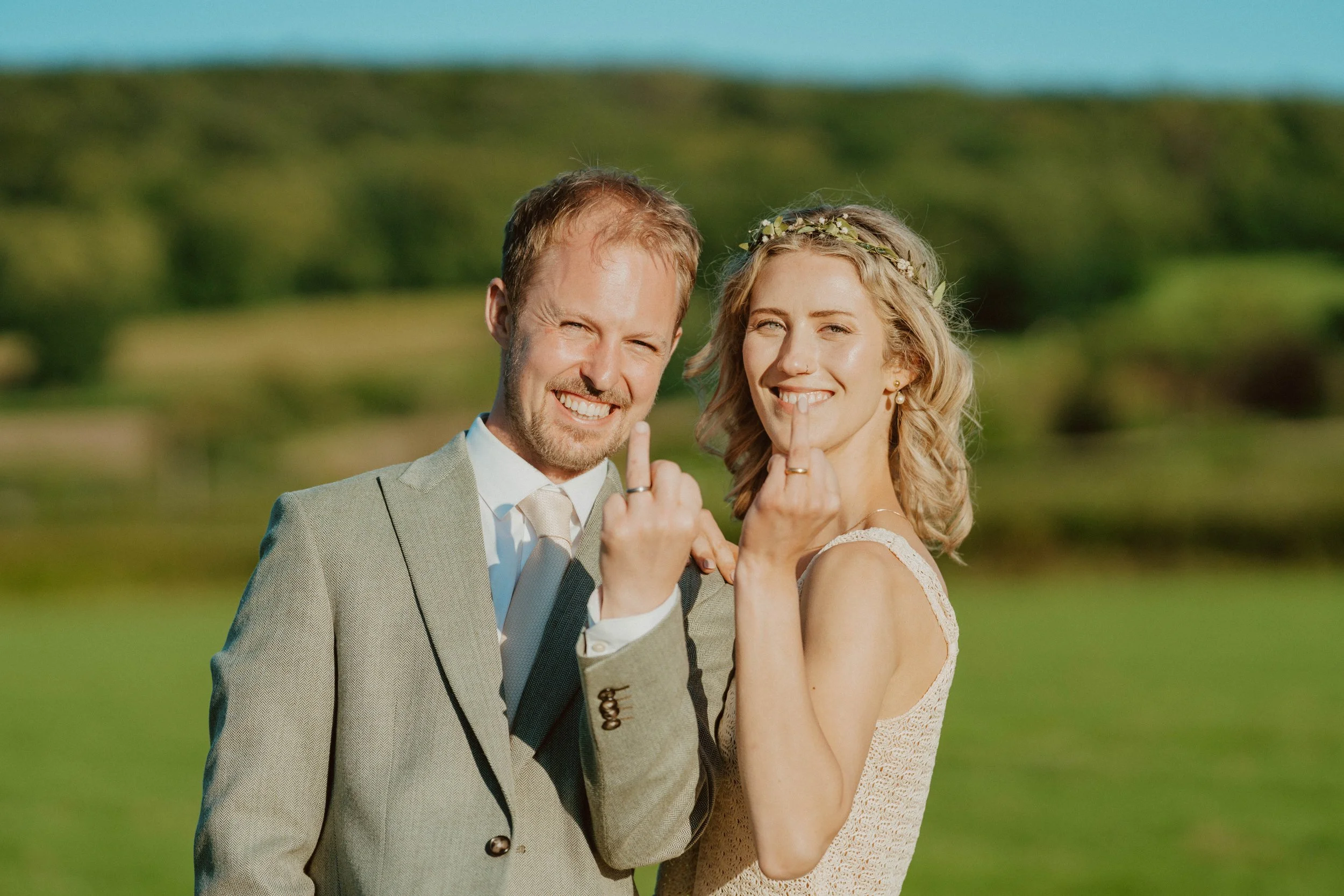 A man and woman in wedding attire showing their middle fingers with smiles, outdoors on a sunny day with a green landscape background.