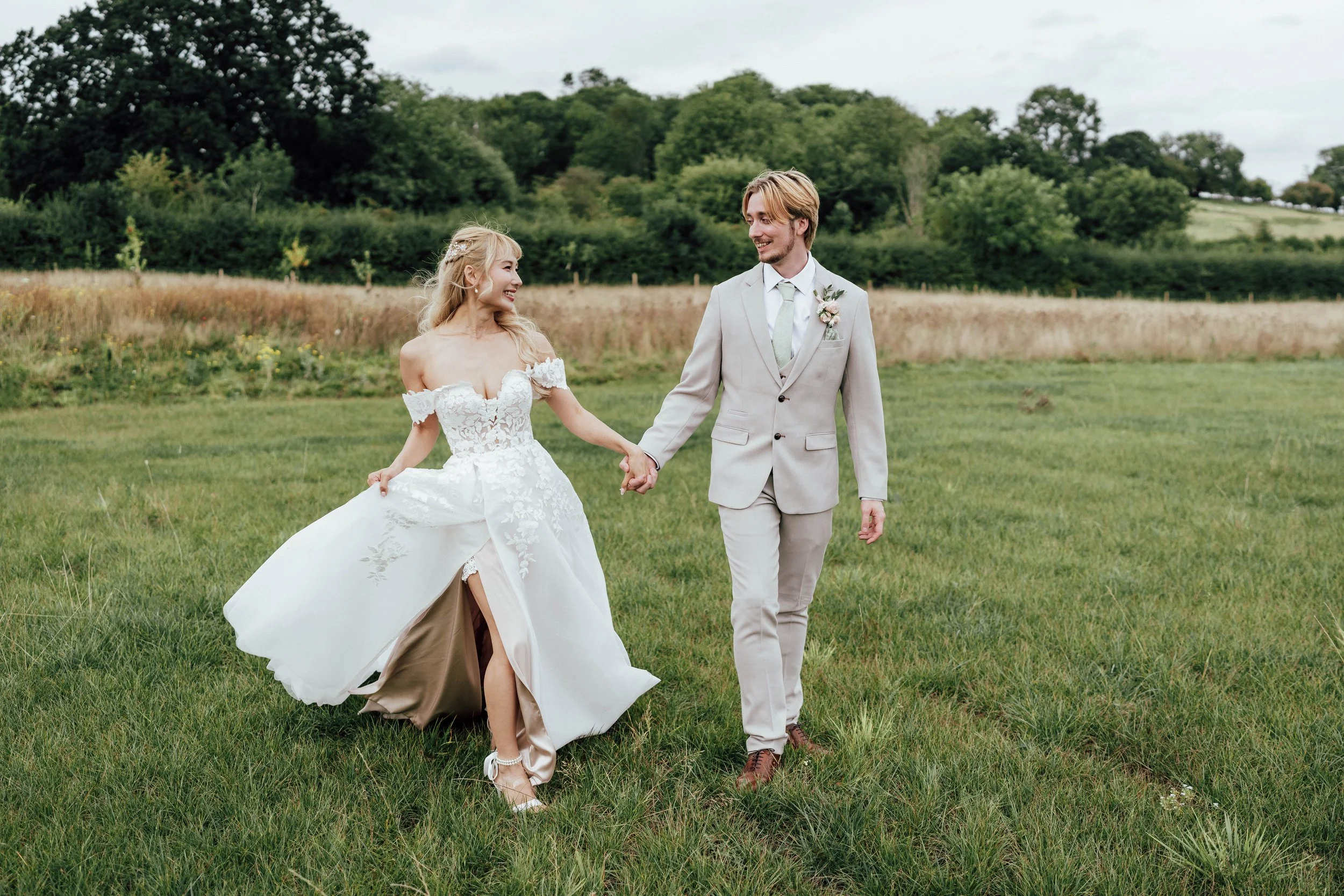 A newlywed couple walking hand in hand on a grassy field, with trees and hills in the background. The bride is in a white lace wedding dress, and the groom is in a light gray suit.