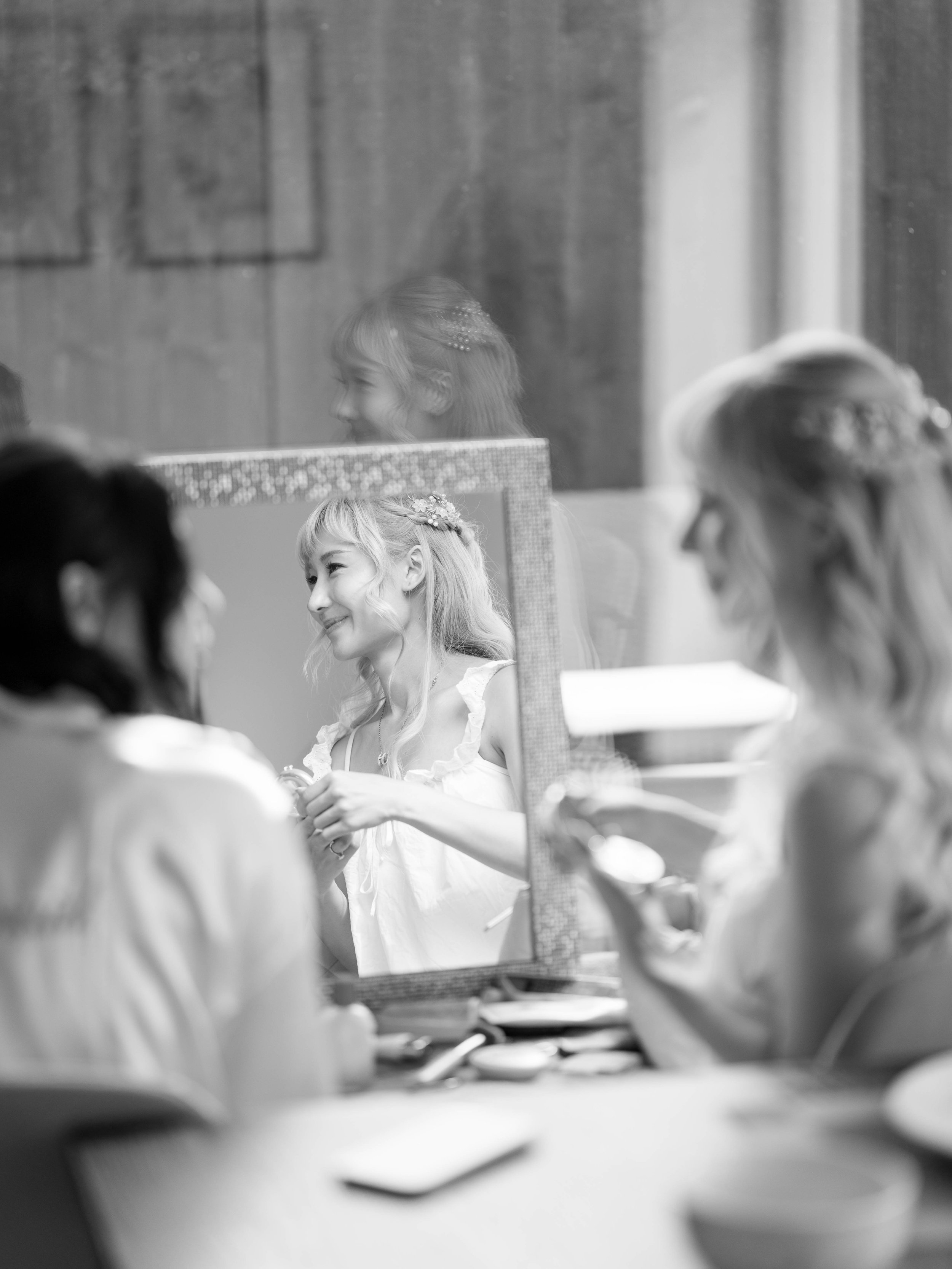 A woman is looking at herself in a mirror while applying makeup or grooming, with two other women sitting at a table nearby, in a room with wooden walls.