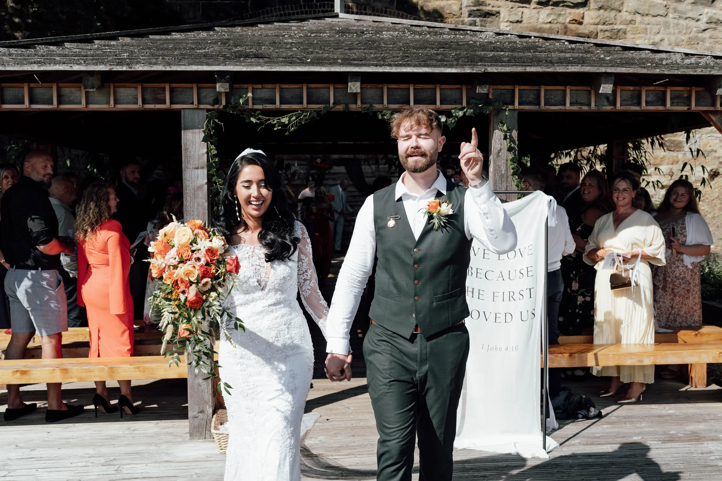 A newly married couple walking hand in hand outdoors, smiling, with guests in the background at a wedding ceremony.