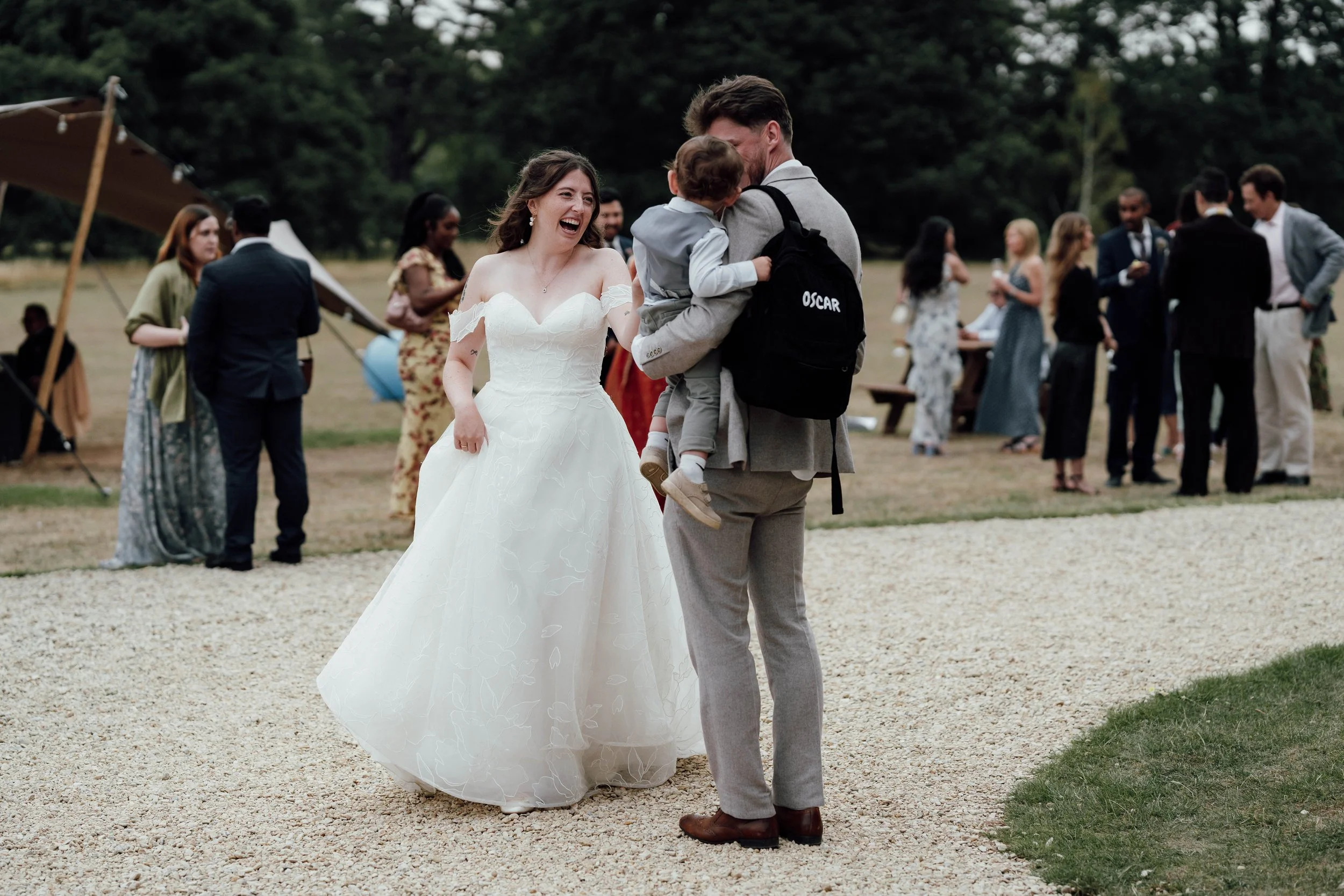 A bride in a white wedding dress laughing while talking to a man holding a child at an outdoor wedding reception, with guests in the background.