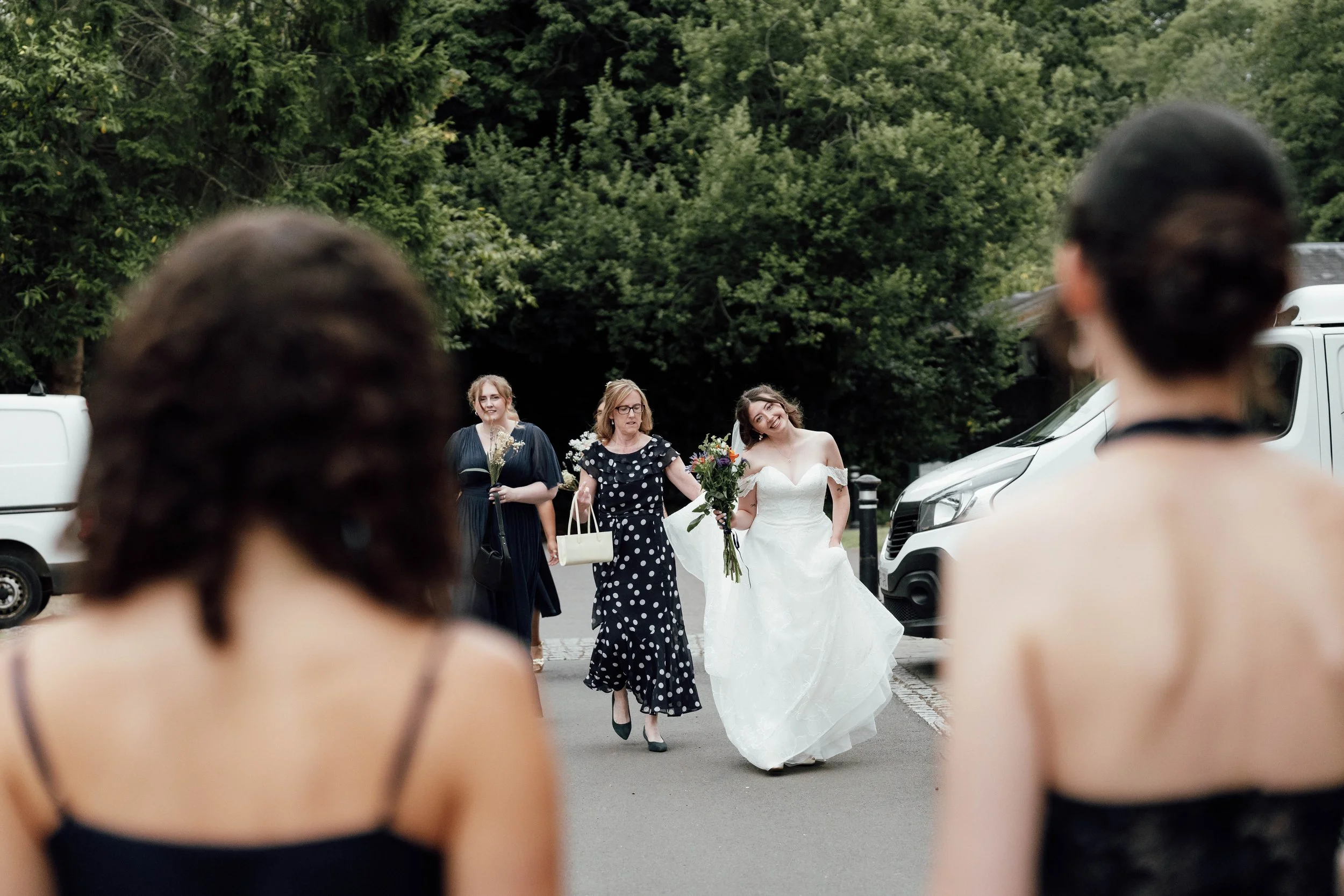 A bride in a white wedding dress holding a bouquet of flowers walking on a street, flanked by two women. Two women are in the foreground with their backs to the camera, one with hair in a bun and the other with short hair, both in black dresses. Cars