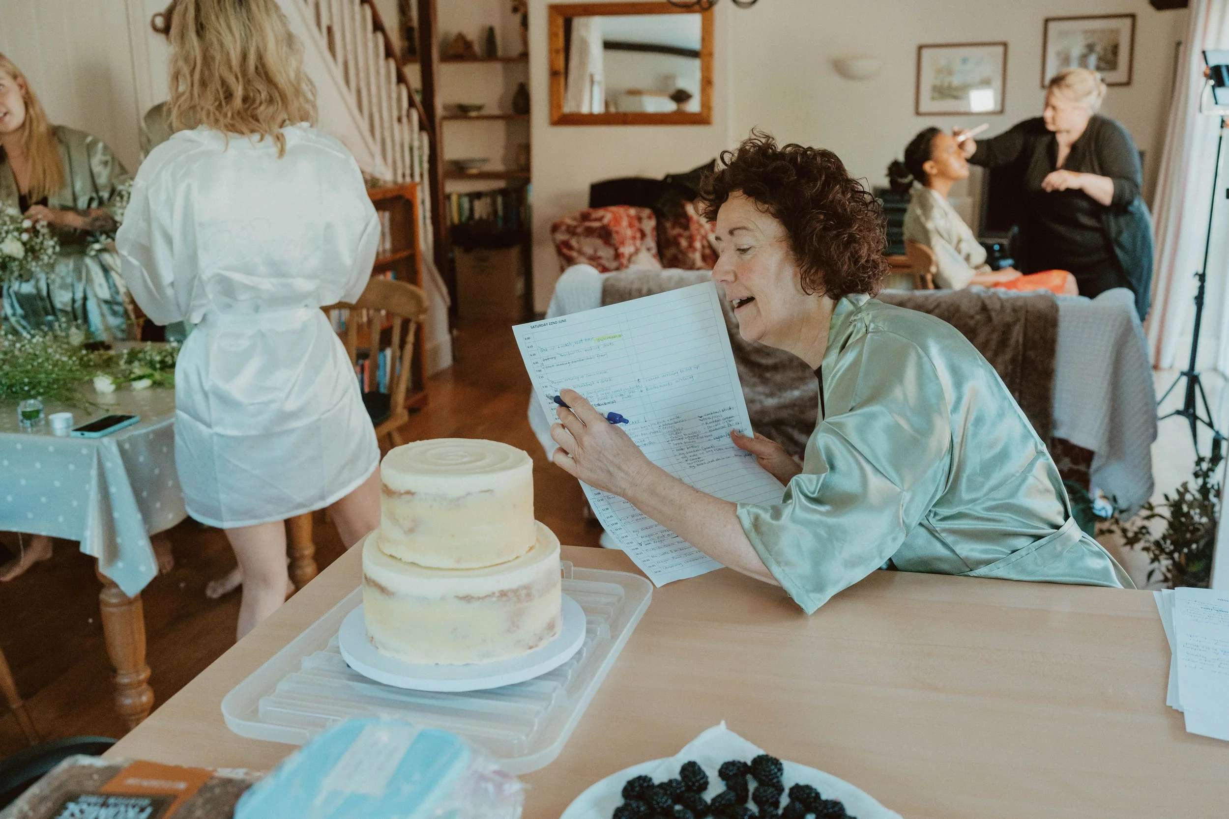 A woman in a mint green satin robe sitting at a table, reading a handwritten note, with two cakes and a bowl of blackberries nearby. In the background, a young woman with long blonde hair and a woman with curly hair in a white satin robe work at a ta