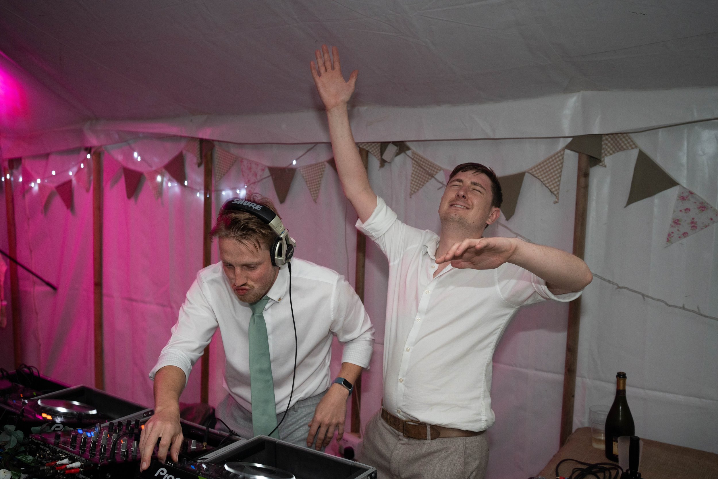 Two men dancing and DJing at a party or event, with decorations and string lights in the background.