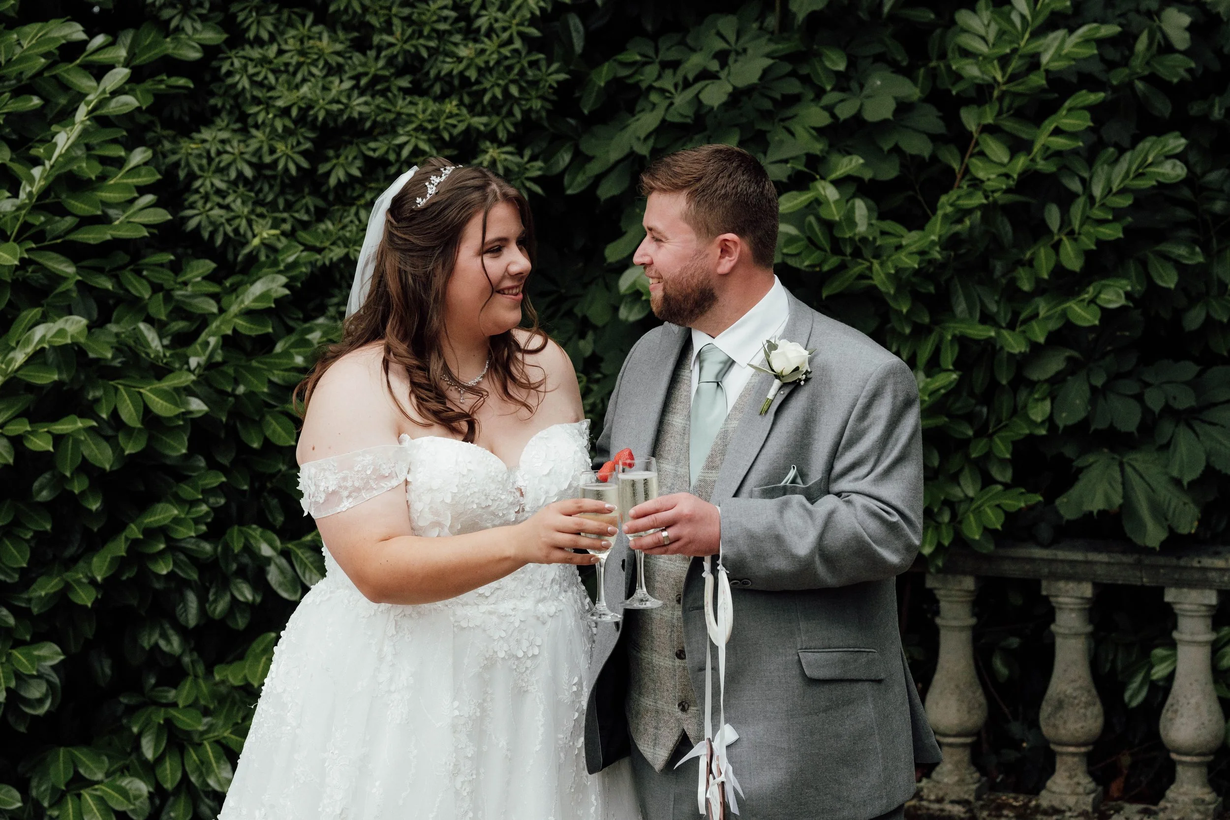 Bride and groom clinking glasses at their wedding celebration outdoors, smiling at each other, with green foliage in the background.